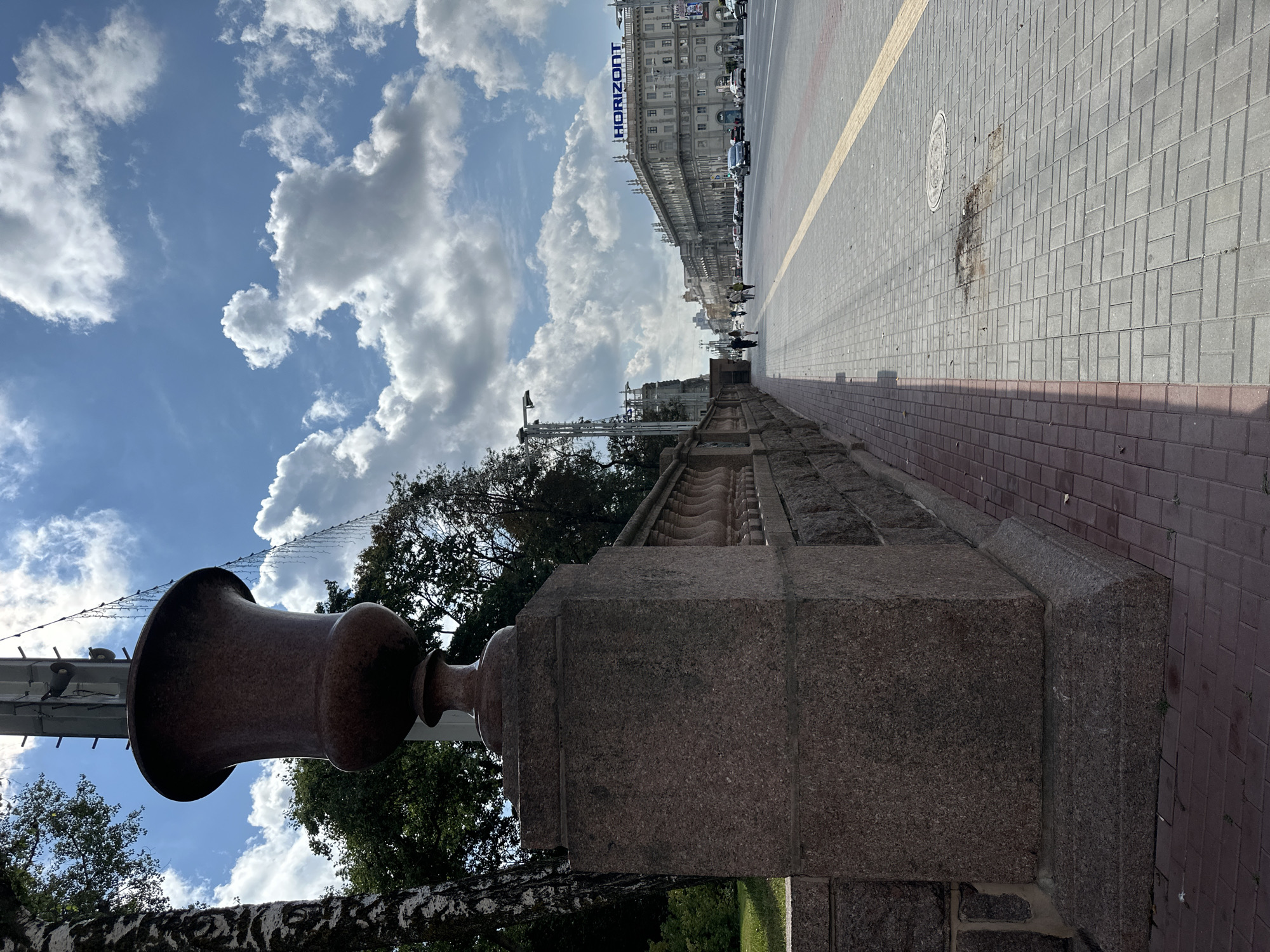The image shows a street scene in Minsk, Belarus, specifically along Independence Avenue. In the foreground, a large, ornate stone planter is visible, set on a granite pedestal. A stone wall with decorative balustrades runs alongside the sidewalk. The sidewalk itself is paved with light gray interlocking bricks, bordered by red bricks. Further down the street, several cars are driving in the same direction, and a few pedestrians are walking on the sidewalk. Buildings line the opposite side of the avenue, with the "HORIZONT" sign prominently displayed on one of them. The sky is blue with scattered white clouds, and sunlight casts shadows on the pavement.