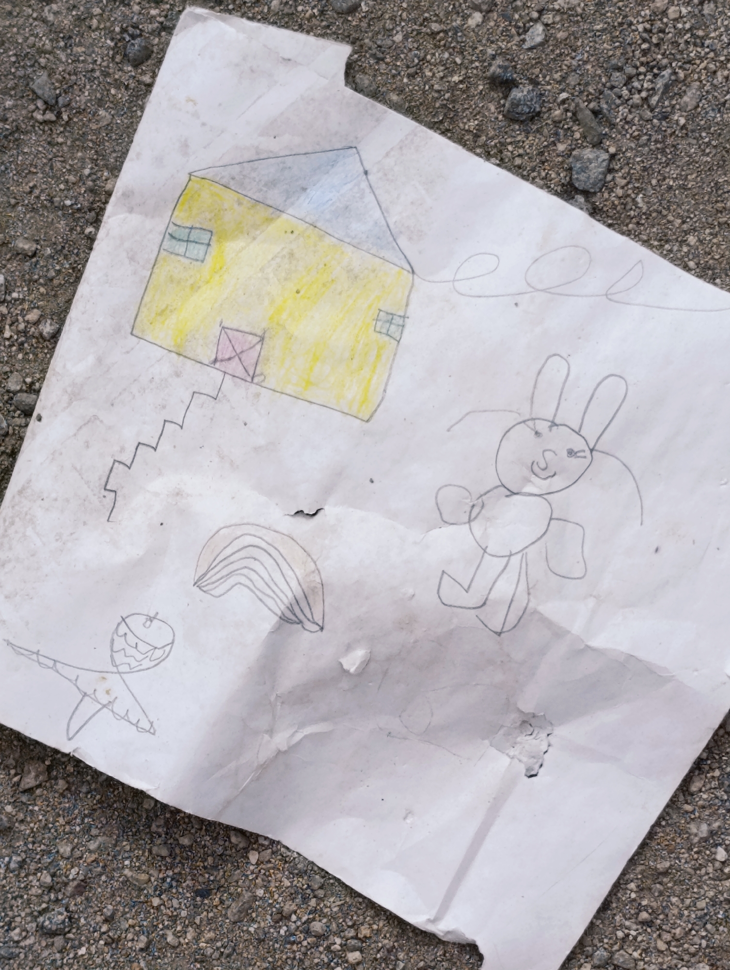 The image shows a crumpled piece of paper with a child's drawing on it, lying on a surface of small grey stones and gravel. The drawing depicts a yellow house with a gray roof, two windows, and a door with a red cross. A set of stairs leads up to the door. To the right of the house is a drawing of a bunny with long ears, round eyes, and a smiling mouth, with two small arms and legs. Below the house and to the left, there is a drawing of an acorn or a pinecone with a small stem and what appears to be a leaf attached. A wavy line with a swirl is drawn above the bunny. The paper is dirty and has a small hole in the lower right section. The background is a natural, outdoor environment, typical of a street or path in the city of Leh, India, characterized by the prevalent use of stones and gravel for surfaces.