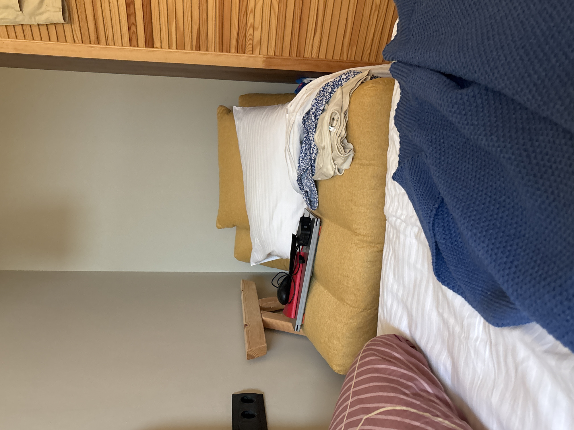 The image depicts a cluttered bed in a room, possibly a bedroom or a studio apartment. The bed is covered with a white striped duvet. On the bed, there is a yellow futon-style mattress with a white pillow and a pile of folded clothes, including a blue and white patterned garment. To the left of the bed, a pink and brown striped pillow rests on the duvet. In the background, a wooden cabinet with slatted doors is visible on the right. A black electrical outlet is mounted on the wall to the left. Resting on the yellow futon is a laptop, a red notebook, a computer mouse, and a pair of glasses. A small wooden stool-like object with notches is placed behind the laptop. The scene appears to be in a private dwelling.
