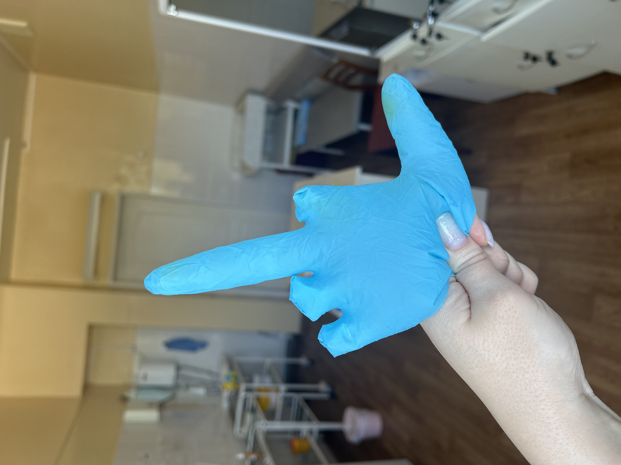 A human hand covered with a blue surgical glove is held up in front of a blurred background of what appears to be a medical or dental office. The glove is shaped to resemble a gun, with the index finger extended forward and the thumb raised. The hand belongs to a person with fair skin and glittery nail polish on at least one visible fingernail. The background includes medical equipment and furniture, and the color scheme of the room consists of light yellow and brown tones. The specific location of Фоки, Russia cannot be determined from the image alone.