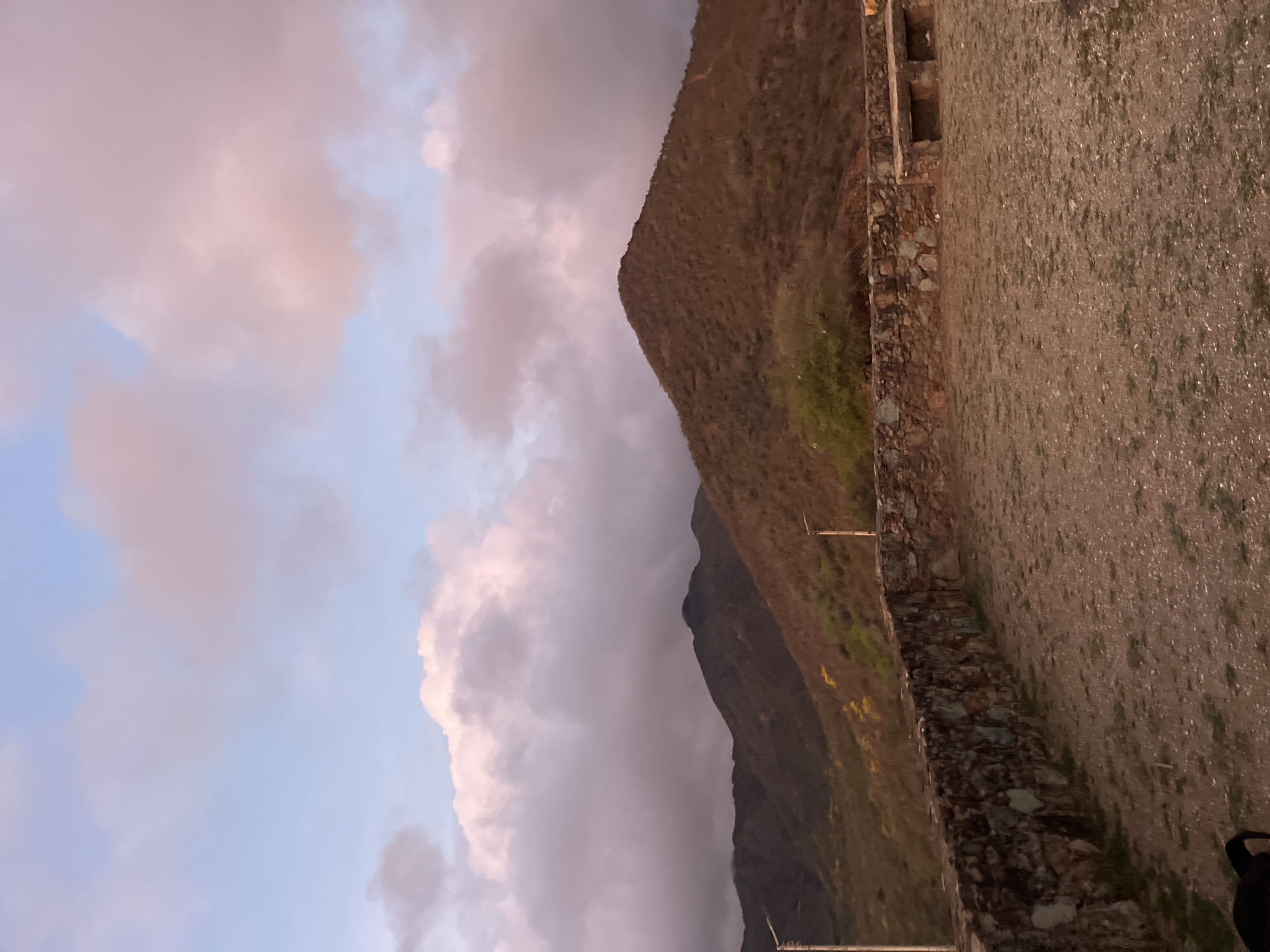 The image depicts a mountainous landscape at sunset. The sky is filled with soft pink and purple clouds against a pale blue backdrop. In the foreground, there is a low stone wall bordering a gravelly area with some sparse vegetation. Behind the wall, the mountains are covered in dry, brown brush, with hints of yellow wildflowers in the distance. Two lampposts are visible on the slopes. The scene is tranquil and evokes a sense of natural beauty. The location is identified as Santa Ana, Venezuela.