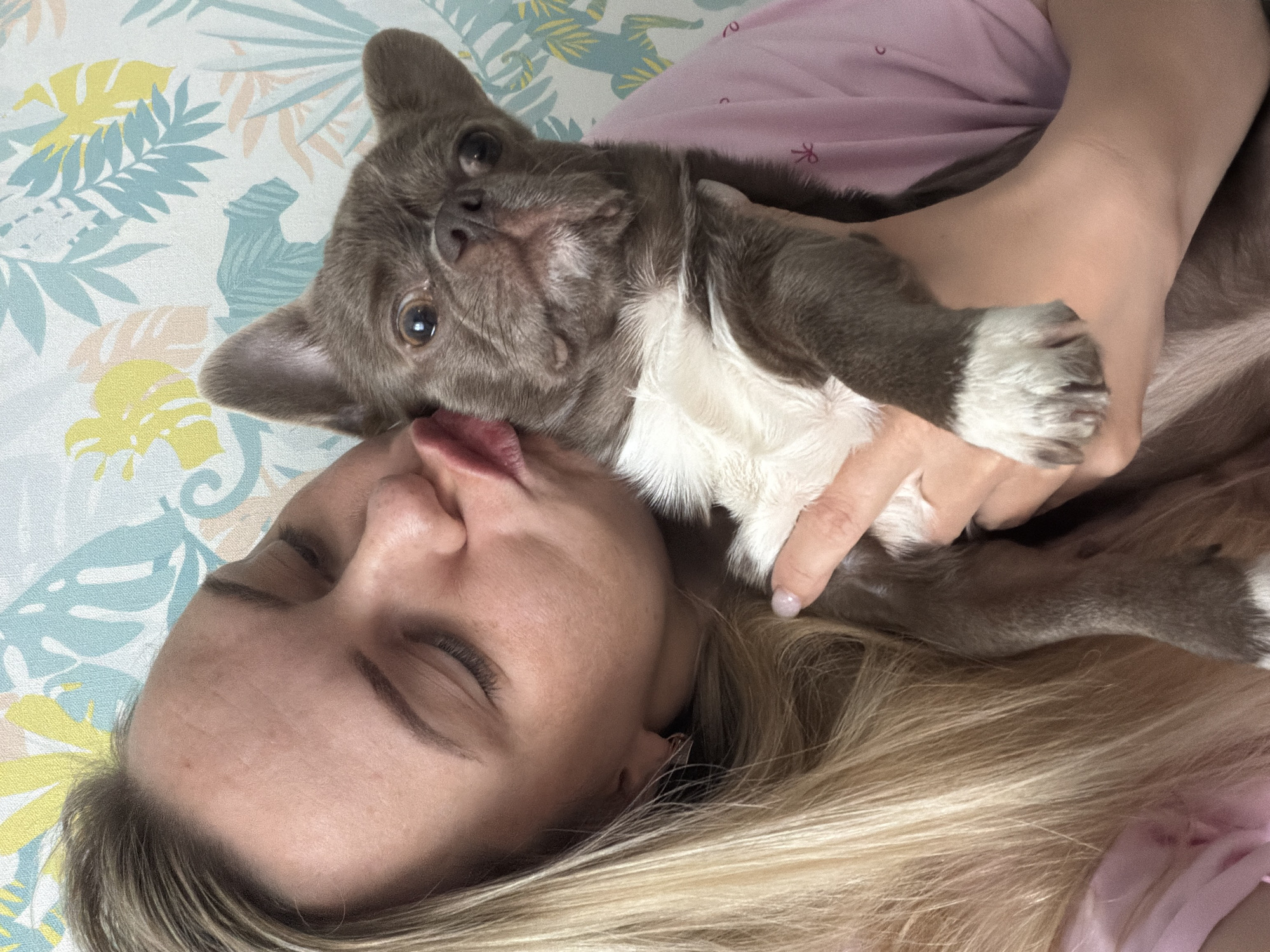 A woman with blonde hair is lying down with her eyes closed, kissing a gray and white bulldog. The woman is wearing a pink shirt. They are lying on a bed with a floral patterned sheet. The photo does not appear to have been taken in the city of Votkinsk, Russia.