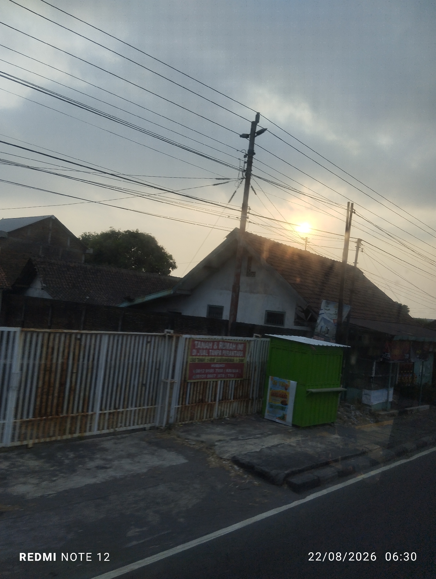 The image shows a street scene in Kertonatan, Indonesia, at sunrise. The sky is overcast with the sun appearing as a bright orb peeking through the clouds. Overhead, a complex network of power lines crisscrosses the sky, supported by utility poles. In the foreground, a rusty metal fence runs horizontally across the frame, partially obscuring a row of buildings with tiled roofs. To the right of the fence, a bright green, boxy structure stands beside a building. A sign on the fence is partially visible, featuring text in Indonesian. The street in front of the buildings appears to be paved with asphalt, with a white line marking the edge. The overall impression is one of a residential or commercial area on the outskirts of a town, captured during the early morning hours. A watermark in the bottom left corner reads "REDMI NOTE 12" and in the bottom right corner "22/08/2026 06:30".