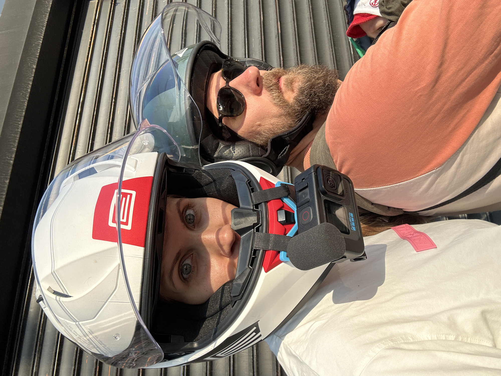 Two individuals, a woman and a man, are pictured from the chest up, both wearing motorcycle helmets and facing forward, positioned side-by-side. The woman, in the foreground on the left, has her eyes visible through a clear visor and is wearing a white helmet with red and black accents. A black GoPro camera is attached to her helmet with a mount. She is wearing a white t-shirt with a pink rectangular patch. The man, to her right and slightly behind, wears a dark green helmet and dark sunglasses. He has a beard and is wearing a peach-colored t-shirt. They appear to be outdoors, with a textured metal roller shutter door in the background. The lighting suggests it is daytime, casting shadows on the subjects. The scene is set in Fethiye, Turkey, likely on a street or in an urban area.