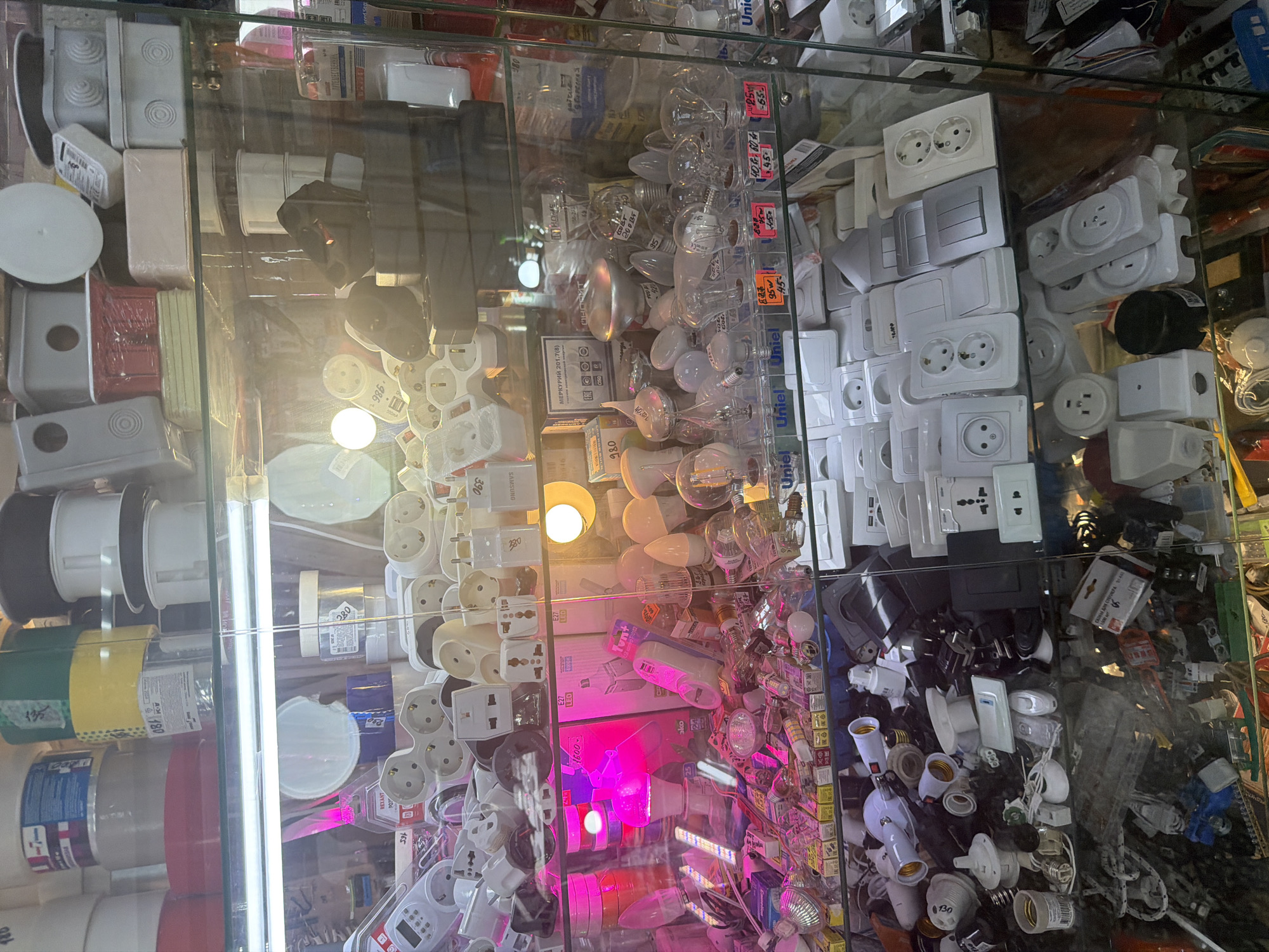 This is a well-lit, eye-level, medium shot of a retail display inside an electrical supply store. The store is located in the city of Tyumen, Russia. The display features a wide variety of electrical components and lighting fixtures. The main items visible are light bulbs, electrical outlets, switches, adapters, and junction boxes. The items are neatly arranged on glass shelves within a refrigerated display case. The lighting in the display case is a mix of cool white and pink hues, with some brighter spotlights illuminating specific areas. The overall impression is one of an organized and well-stocked store, catering to a wide range of electrical needs. There are no people visible in the image.