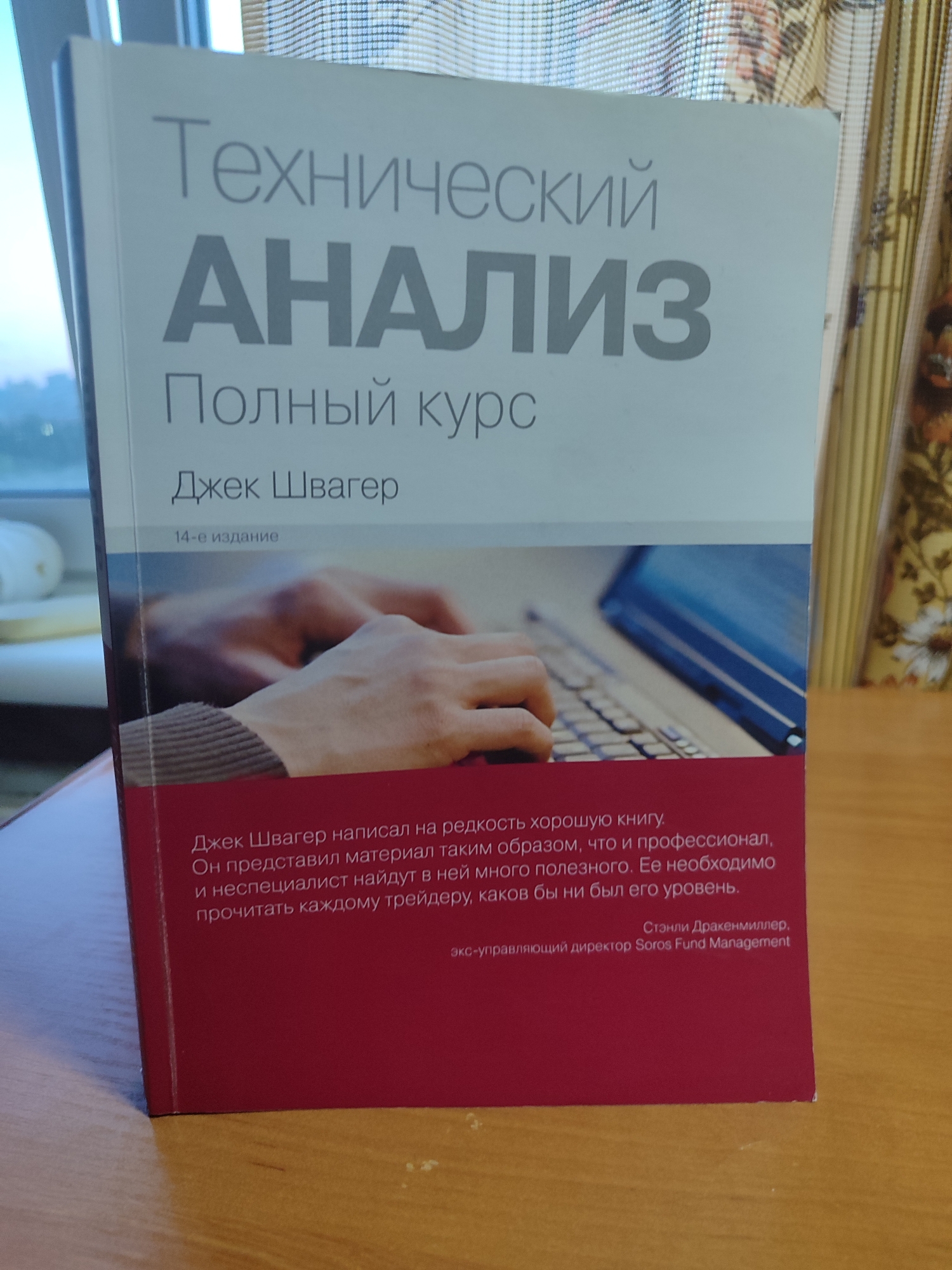 The image shows a book with a red and white cover, titled "Технический АНАЛИЗ Полный курс" which translates to "Technical ANALYSIS Complete Course". The author's name, "Джек Швагер" (Jack Schwager), is also visible. Below the title and author, it states "14-е издание" (14th edition). The cover features a photograph of hands typing on a laptop. The bottom portion of the cover contains text, likely a blurb or review, in Russian. The book is placed on a wooden surface, possibly a table, with a blurred background suggesting an indoor setting with a window and curtains. The lighting suggests it might be daytime.