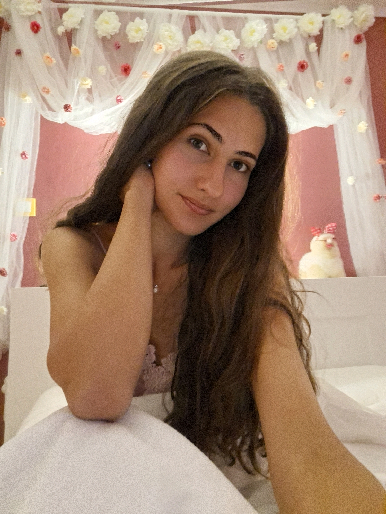 A young woman with long, brown, wavy hair is in bed. She is wearing a pink lace camisole and a pearl necklace. Her right arm is bent, and her hand is behind her head. She is looking at the camera with a slight smile. The background is a pink wall with sheer white fabric draped from the ceiling, decorated with artificial flowers. A small stuffed chick with a red bow is on a shelf behind her. The scene appears to be in a bedroom.