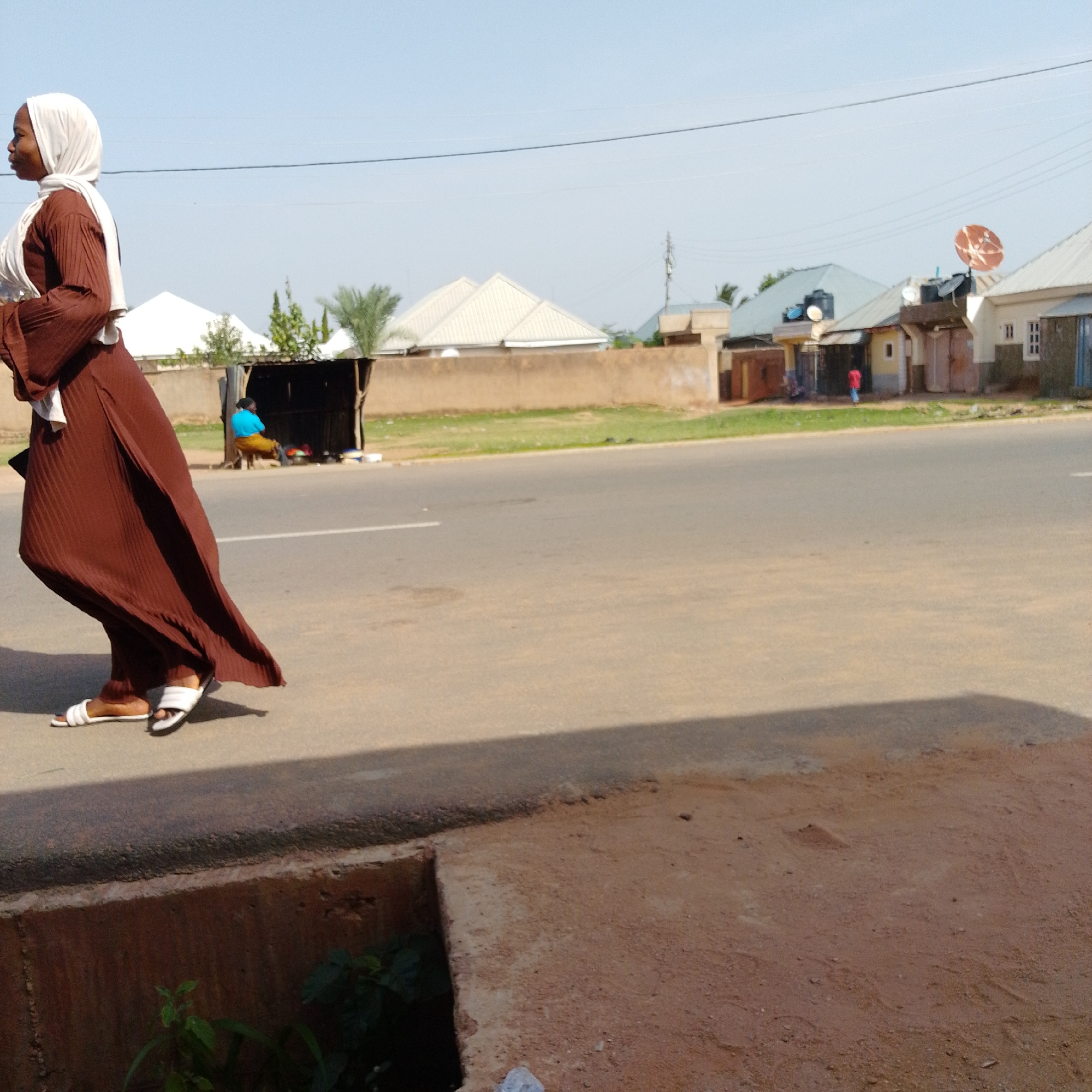 In Gombe, Nigeria, a woman wearing a brown dress and white hijab walks along a dusty road. She is on the left side of the image, with her body angled towards the right. The road is paved and appears to be a main thoroughfare, with a white line marking the lane. In the background, several buildings with corrugated metal roofs are visible, typical of the architecture in Gombe. A few palm trees and other vegetation dot the landscape. Further in the distance, utility poles and wires indicate the presence of infrastructure. The sky is clear and a pale blue, suggesting a sunny day. Another person is visible sitting under a makeshift shelter on the right side of the road, wearing a blue top and yellow skirt.