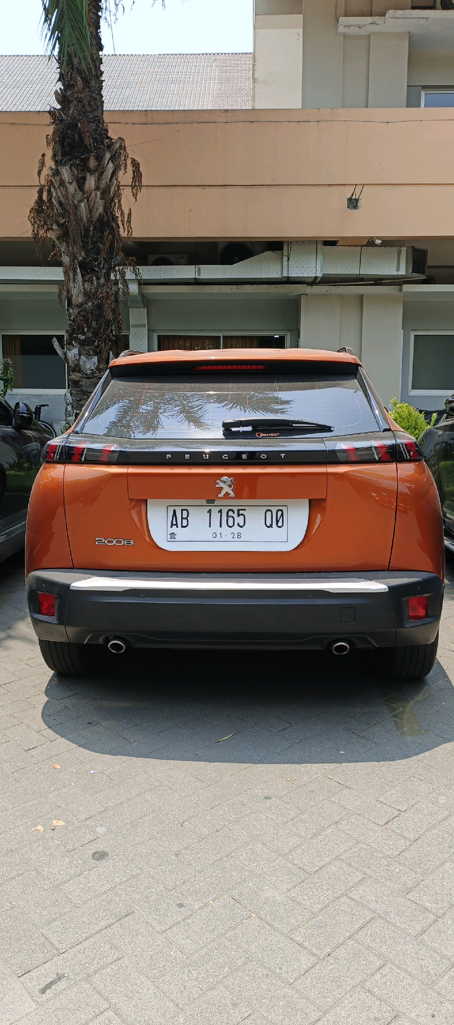 A rear view of an orange Peugeot 2008 is parked on a herringbone pattern brick paved surface in front of a light brown building. The car has a black trim around the rear window, with the "PEUGEOT" logo in the center. A license plate with the characters "AB 1165 QQ" and the date "01 . 28" is mounted on the rear. The car also has dual exhaust pipes. A palm tree is visible to the left of the car, and part of another dark-colored vehicle is to the left. The building has several windows with dark blinds. The image appears to have been taken in Ambarketawang, Indonesia.