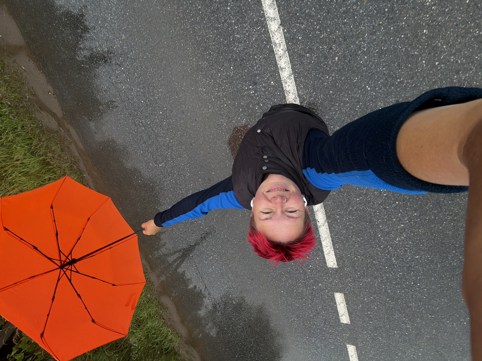 A person with bright pink hair and a nose ring, wearing a black vest over a blue and black long-sleeved shirt, smiles as they hold up a bright orange umbrella while standing on a wet asphalt road. The road has a white dashed line and a solid white line. The environment appears to be a roadside in Chaikovsky, Russia, with green grass visible on the left edge of the frame. The scene is captured from a high angle, likely a selfie.