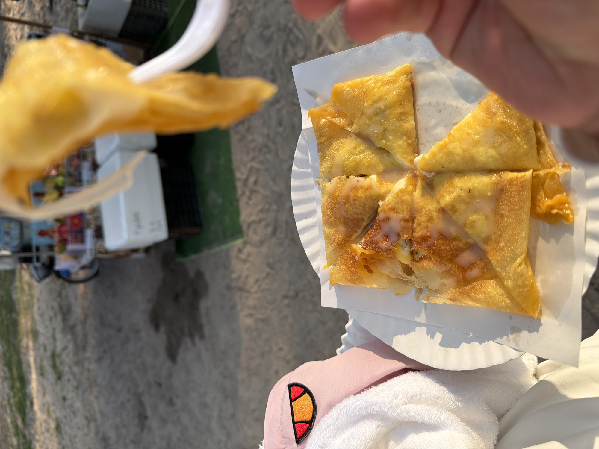 A golden-brown flatbread, likely a Thai Roti, cut into triangular sections and drizzled with a white glaze, is presented on a white paper plate. A single piece of the flatbread is actively being lifted from the plate by a white plastic fork held by an unseen individual's fingers on the right side of the frame. To the left, a portion of a pink plush item with an orange and red patch (resembling an eye) is visible, resting against a white towel or blanket. The scene is outdoors, with a sandy or dirt ground featuring dark, irregular wet patches. In the blurred background, a vendor's cart is discernible, displaying colorful items, possibly fruit, and a white cooler unit, indicating an informal market or street food environment in Choeng Thale, Thailand.
