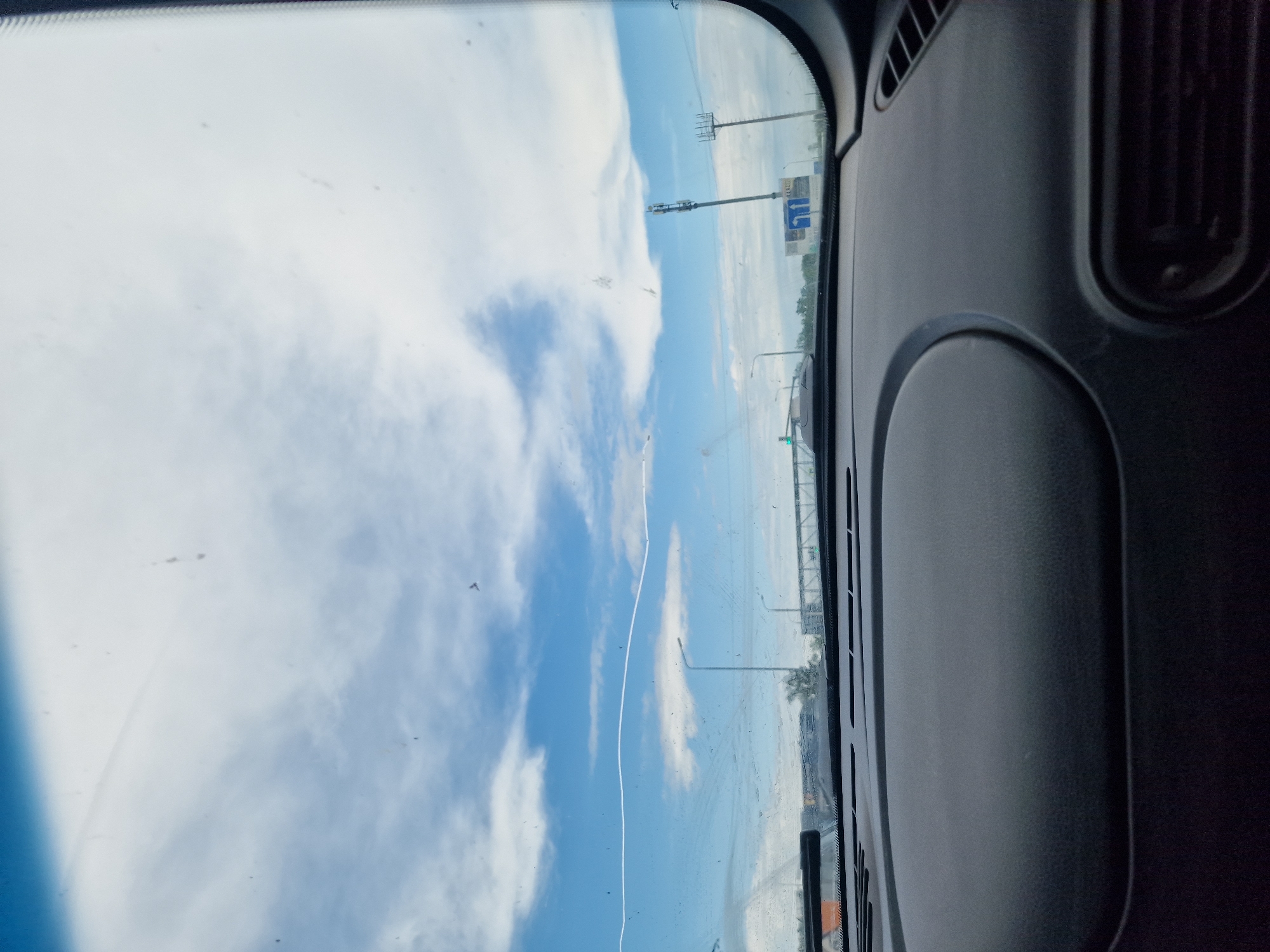The image is taken from inside a vehicle looking out through the windshield. The view shows a bright blue sky with scattered white clouds. A thin, white horizontal line, possibly a contrail from an airplane, is visible across the sky. In the distance, there are road signs, streetlights, and cellular towers. The dashboard of the vehicle is visible in the foreground, appearing dark and textured. The overall scene suggests a daytime drive on a highway or a similar road. There is no specific indication of the location being Zmeika, Russia, or any other city.