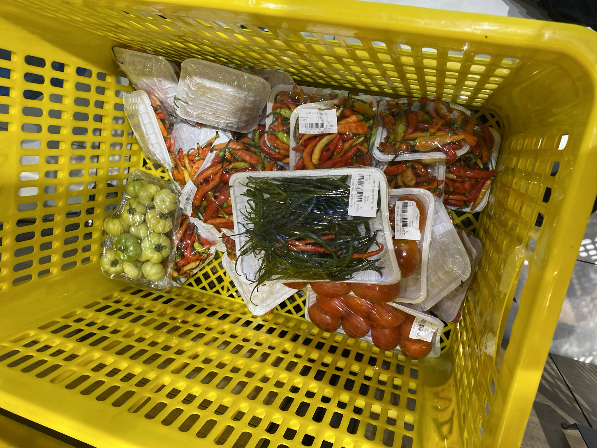 A bright yellow plastic shopping basket is filled with various produce. The basket is seen from a high angle, looking down into its contents. The produce includes bunches of green tomatoes, several packages of red and green chili peppers of varying sizes and heat levels, and at least one package of tomatoes. The items are mostly contained within clear plastic packaging, with some labels visible, though they are not legible in detail. The setting appears to be a market or grocery store, as suggested by the basket and the organized presentation of produce. The visible part of the basket is clean and shows no signs of damage. The lighting in the scene is bright, indicating an indoor environment.