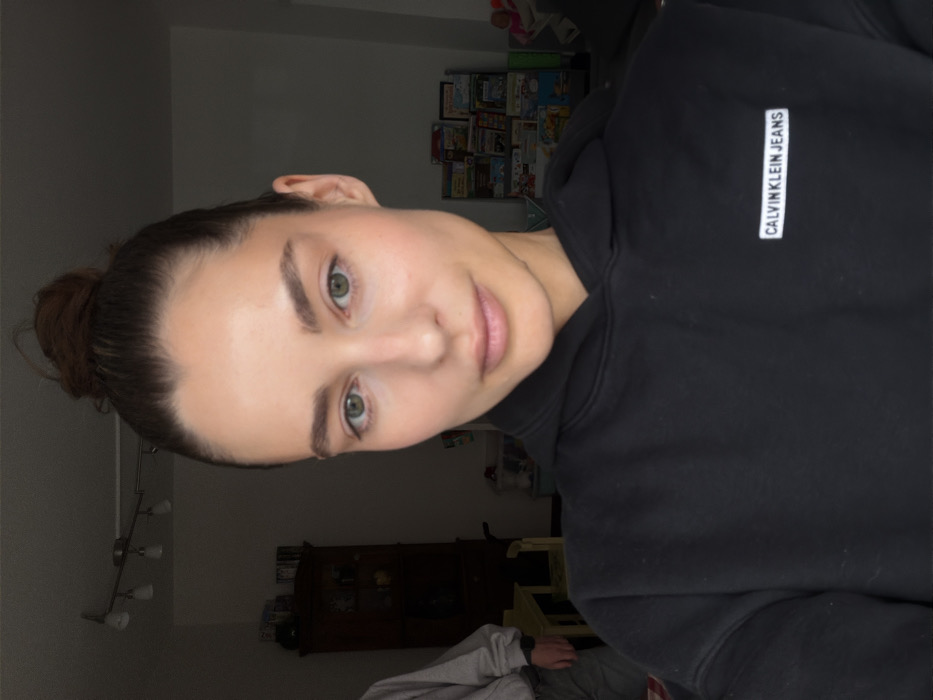 A forensic description of the scene in Unknown, Unknown:
The primary subject is an adult female positioned in the foreground, directly facing the camera. She has light skin, green eyes, and dark brown hair pulled into a high bun. She is wearing a black hooded sweatshirt featuring a white rectangular label on the left chest with "CALVIN KLEIN JEANS" printed in black text. Her expression is neutral.
In the background, to the left of the primary subject, the lower torso and arms of a second individual are partially visible, clad in a light grey long-sleeved top and light-colored trousers, with a watch on the left wrist. Adjacent to this individual, a partial view of a yellow wooden children's table is present. Further back on the left, a dark wooden cabinet or hutch containing various items is discernible. Above, a ceiling-mounted track light fixture with multiple light sources is installed. To the right of the primary subject, a shelf unit filled with colorful children's books and magazines is visible, with additional assorted items and toys in the immediate vicinity. The room is an indoor residential space with uneven lighting, where the foreground is brighter than the background.