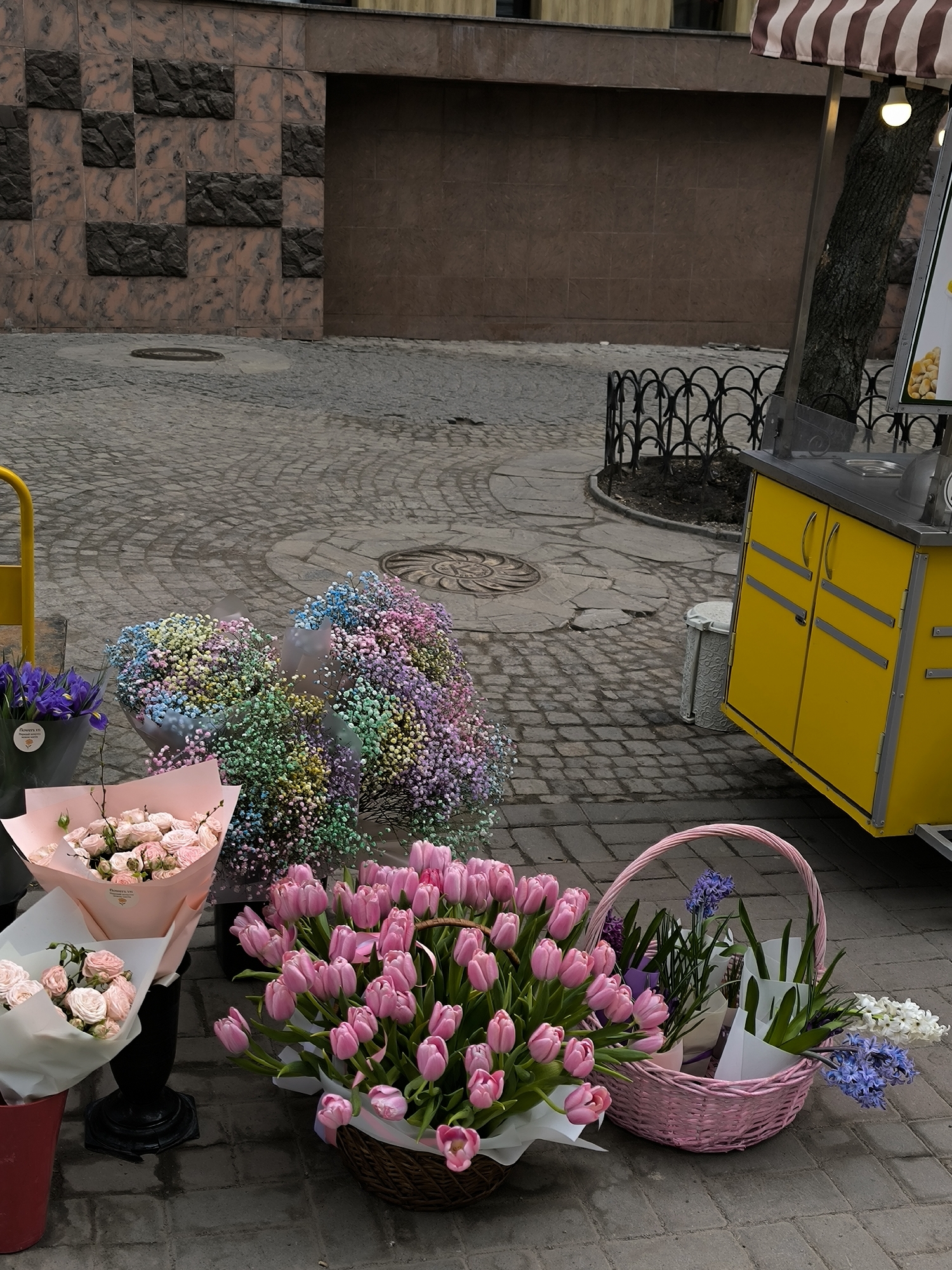 The scene shows a flower stall on a cobblestone street, likely in Vinnytsia, Ukraine, judging by architectural style and typical street furnishings. The main focus is a vibrant array of flowers: bouquets of pink tulips in a wicker basket, arrangements of multicolored gypsophila, and other bunches of roses and irises in wrapping. A pink wicker basket contains flowering plants with white and blue blossoms. A yellow food cart stands nearby, its striped awning partially visible at the top right. The background consists of a building façade constructed with dark stone panels, and a wrought iron fence surrounds a tree. The scene is static and devoid of people.