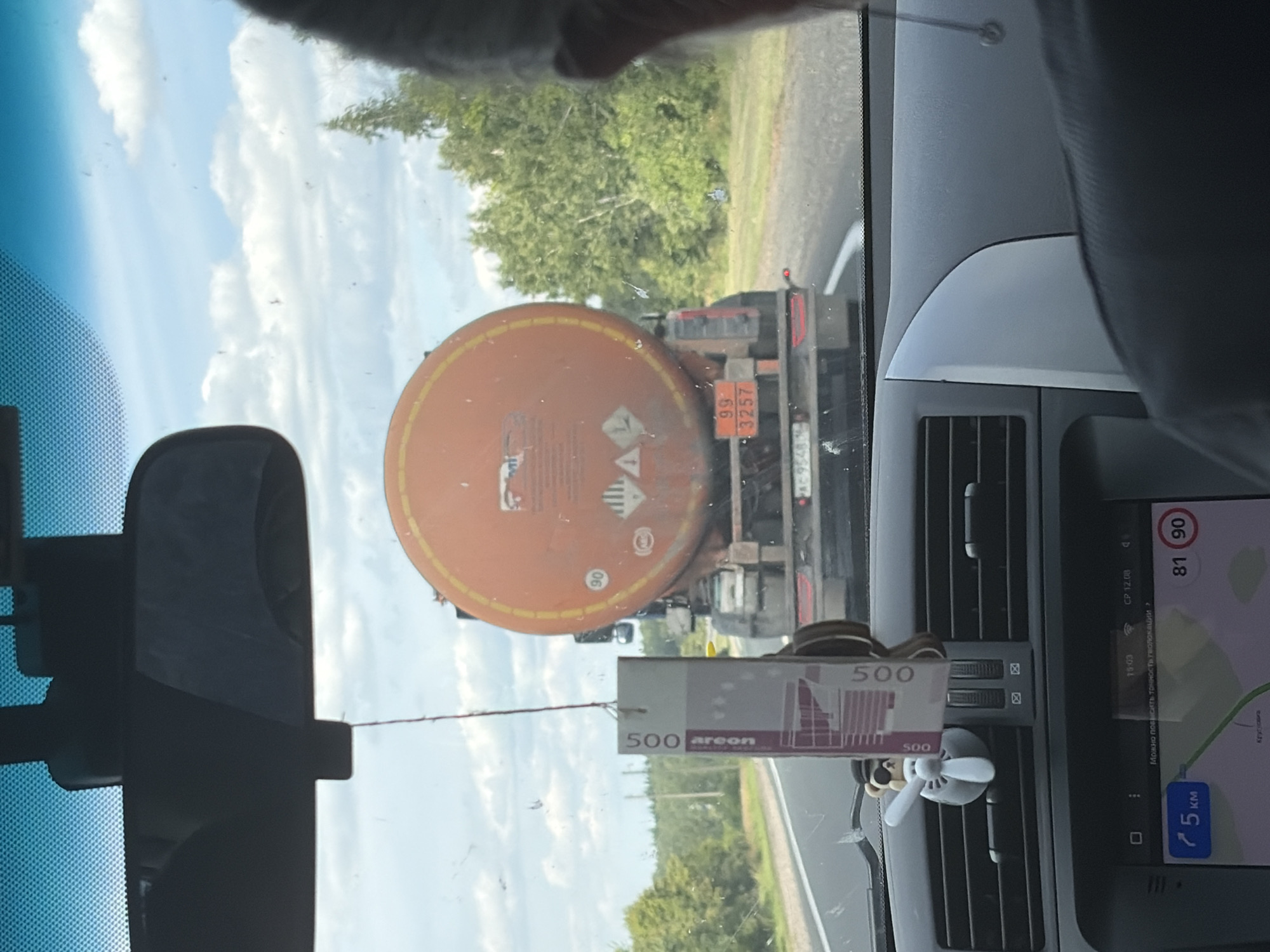 The image shows the interior of a vehicle looking forward through the windshield. In front of the vehicle is a large orange tanker truck. The truck has hazard symbols and numbers "99" and "3257" displayed on the rear. The truck's license plate is partially visible, reading "AC 9548". The road ahead is paved and flanked by trees and vegetation. A car air freshener shaped like a teddy bear with sunglasses and a propeller is hanging from the rearview mirror. On the dashboard, a GPS screen shows a map with the destination labeled as "Kruglovka, Russia" and a distance of "5 km". The time displayed on the GPS is "15:03". The sky is cloudy.