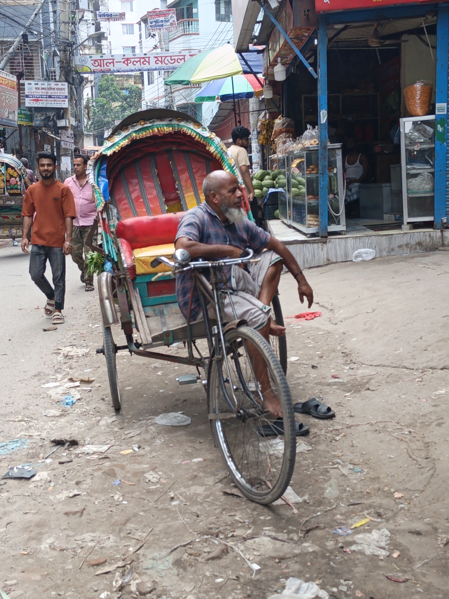The image depicts a street scene in what appears to be a densely populated urban area in Unknown, Unknown. The environment includes buildings with visible signs in Bengali, power lines overhead, and a street littered with debris.
At the center of the scene is a cycle rickshaw, with an older man seated on it. He has a beard and is wearing a checkered shirt and light-colored shorts. The rickshaw is red and green with a simple bicycle frame.
Two younger men are walking along the street. The man on the left is wearing an orange shirt and jeans, while the other is wearing a pink shirt and khaki pants.
To the right of the rickshaw is a small shop with a display of goods, including fruits and baked goods. People are inside the shop, but their actions and details are not clearly visible.
The overall impression is of a bustling street scene in a crowded urban environment.