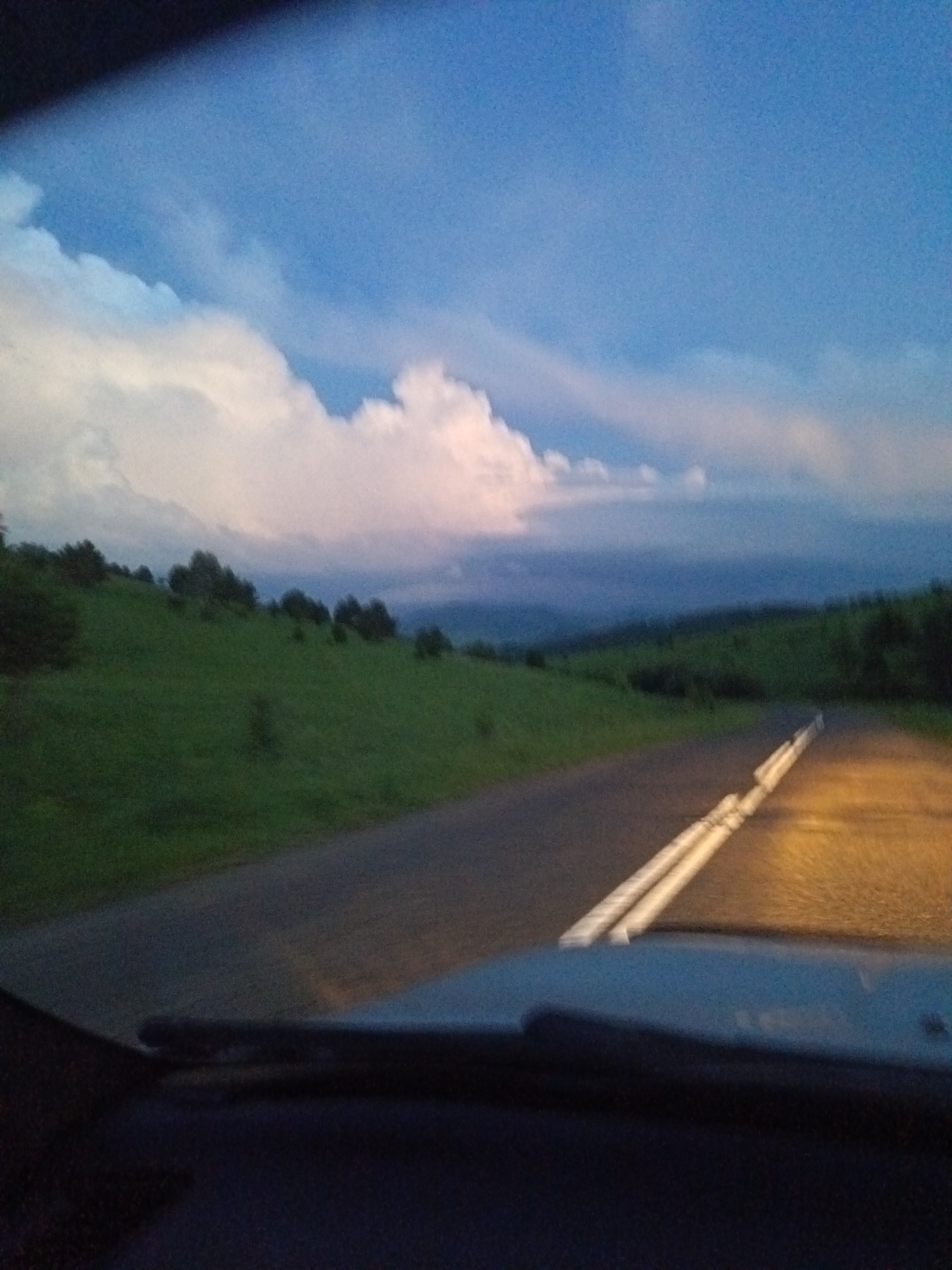 The image depicts a view from a vehicle driving on a paved road, likely at dusk or early evening. The road has white dashed lines marking the lanes. To the left of the road is a grassy hillside dotted with trees. In the distance, rolling hills are visible under a sky with scattered clouds, some of which are illuminated by soft pink and white light. The front of the vehicle's dashboard is visible in the foreground. The location is identified as Sadovyi, Russia.