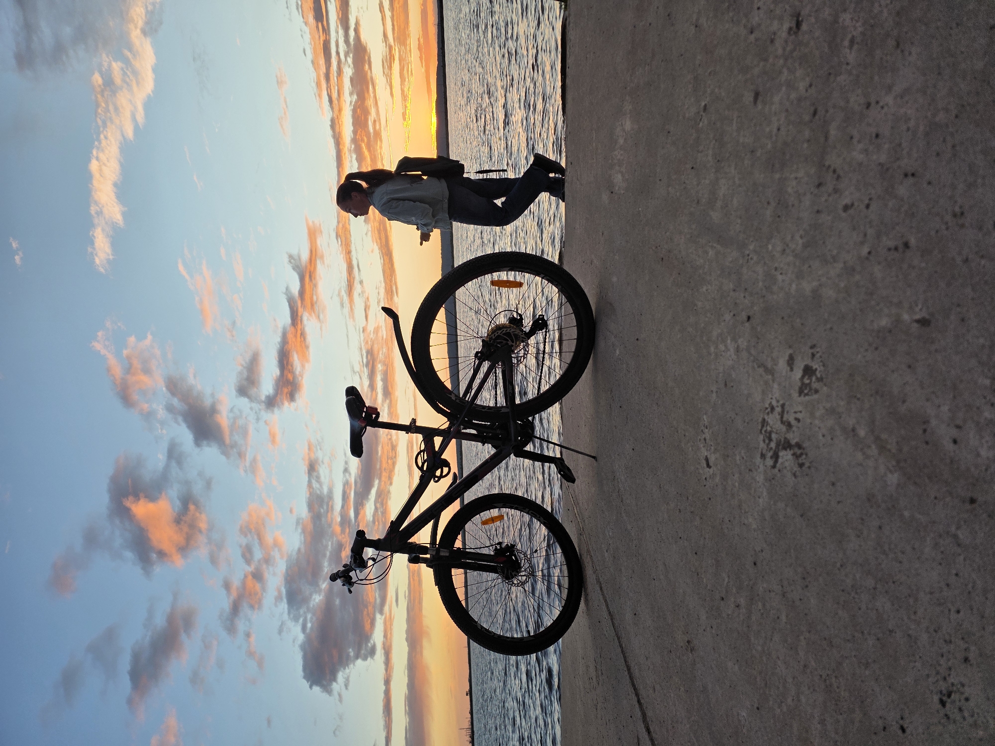 A silhouette of a woman stands on a concrete pier next to a bicycle. She is wearing a light-colored jacket, dark pants, and a backpack. Her hair is pulled back in a ponytail. The bicycle is black and positioned in the foreground, facing left. The background features a body of water, likely a reservoir or river, with gentle waves. The sky is filled with dramatic clouds painted in hues of orange, pink, and blue, indicating a sunset or sunrise. In the far distance, a faint outline of industrial structures can be seen on the horizon, suggesting an urban or industrial setting near Chaikovsky, Russia. The overall scene conveys a sense of peaceful contemplation at dusk or dawn.