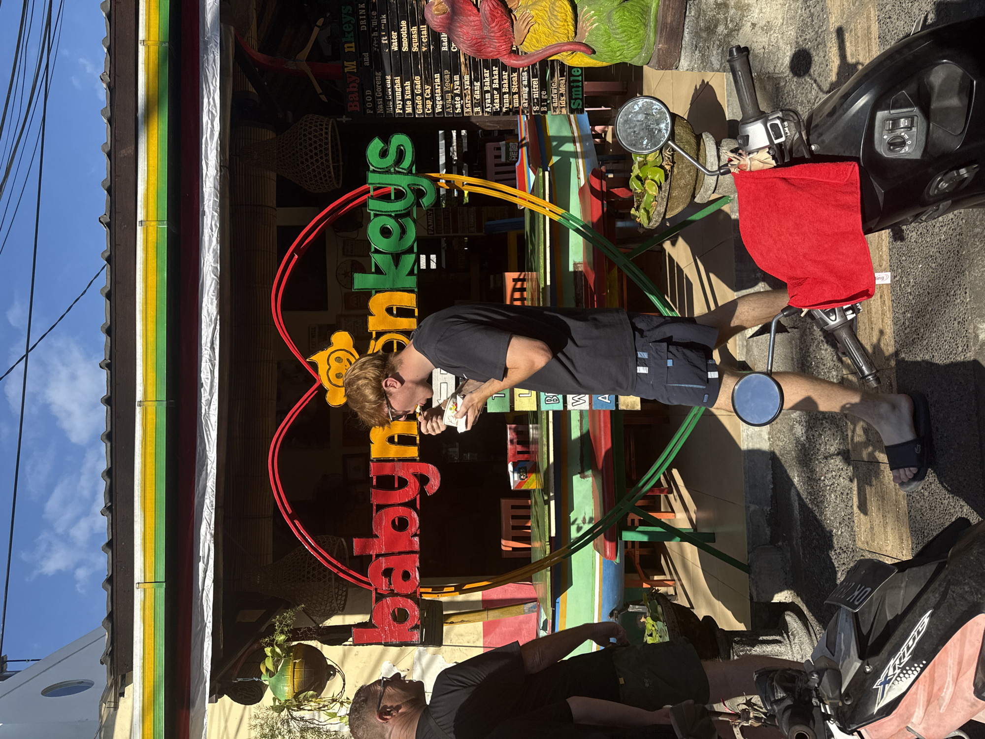 In Denpasar, Indonesia, two men stand on a street in front of a restaurant called "Baby Monkeys." The restaurant has a red neon sign in the shape of a heart with the restaurant's name displayed in red and green lettering. The restaurant has colorful tables and chairs. The man on the left, wearing glasses and a black t-shirt, stands with his back to the camera. The man on the right, with red hair and glasses, is eating food from a white bowl while facing the restaurant. In the foreground, two motorcycles are parked on the sidewalk. The one on the right has a red towel draped over its handlebars. The sky is blue with some clouds.