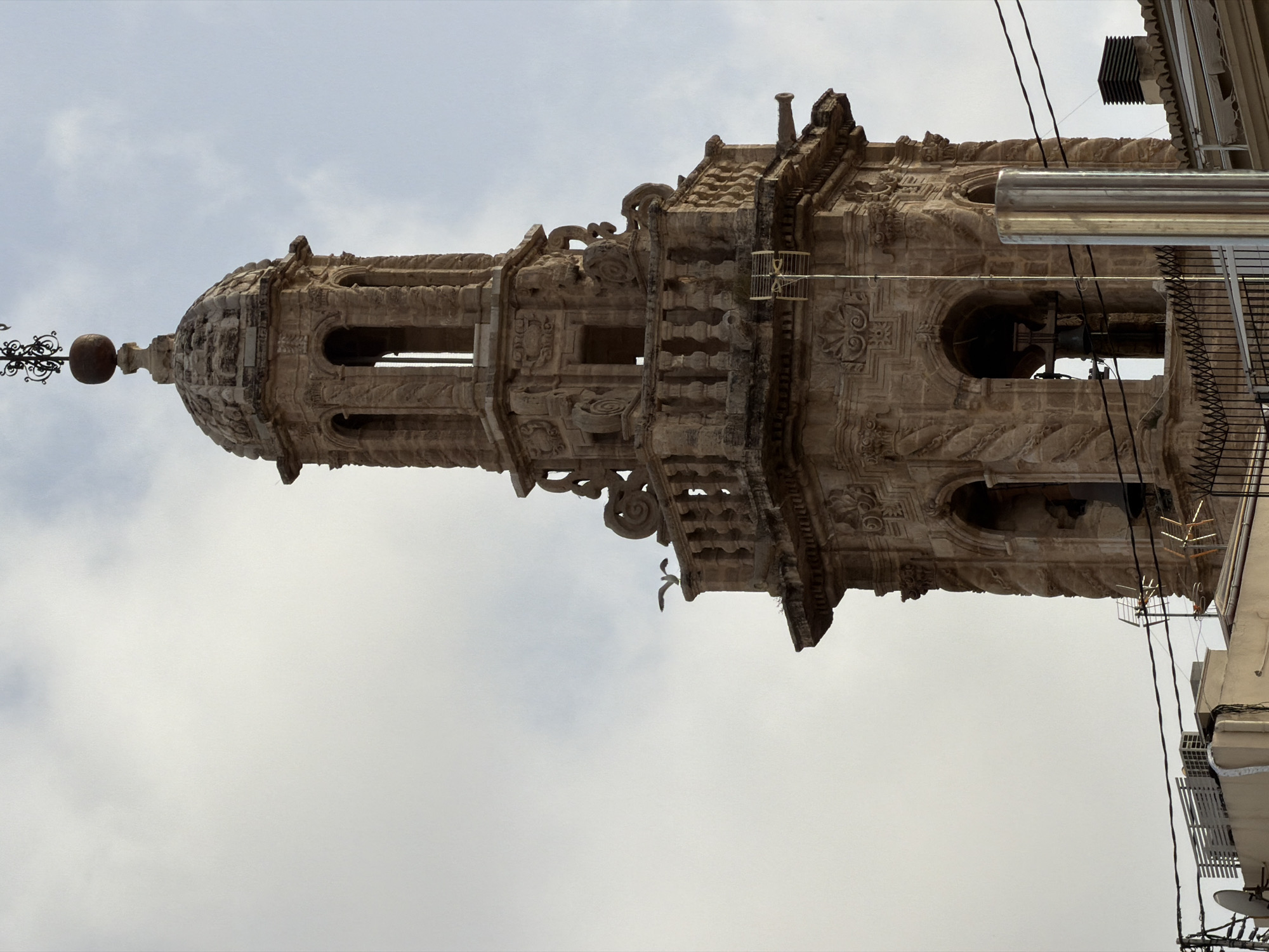This is a waist-up, eye-level, medium shot of a bell tower in a city. The bell tower is made of stone and has a dome-shaped roof. It is decorated with intricate carvings and has several arches. A seagull is perched on the railing of the tower, and the sky is cloudy. In the foreground, there are some electrical wires and part of a building with an air conditioning unit. The scene appears to be an outdoor urban environment.