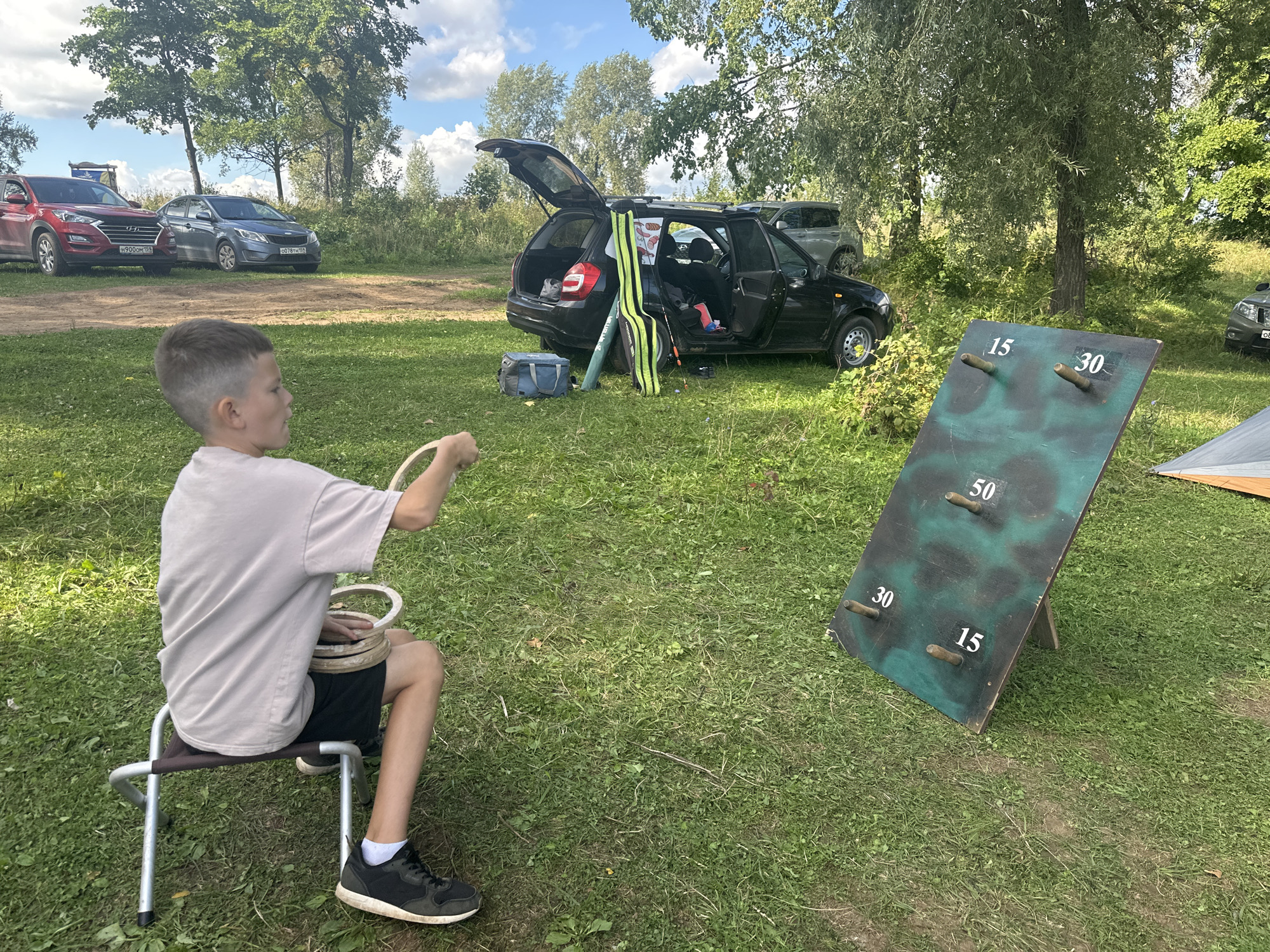 The image depicts a young boy sitting on a folding chair, holding rings, and looking towards a target board in a park-like setting. The target board, which has a camouflage pattern and numbers such as 15, 30, and 50, is propped up on the grass. Several cars are parked in the background, including a black hatchback with its trunk open, revealing camping gear and fishing equipment. The location appears to be a recreational area, possibly a campsite or picnic spot, in Chaikovsky, Russia, given the presence of Russian license plates on the vehicles. The environment is characterized by lush green grass, trees, and a partly cloudy sky. The boy is dressed in a light-colored t-shirt and dark shorts, with athletic shoes. The overall scene suggests a casual outdoor activity or gathering.