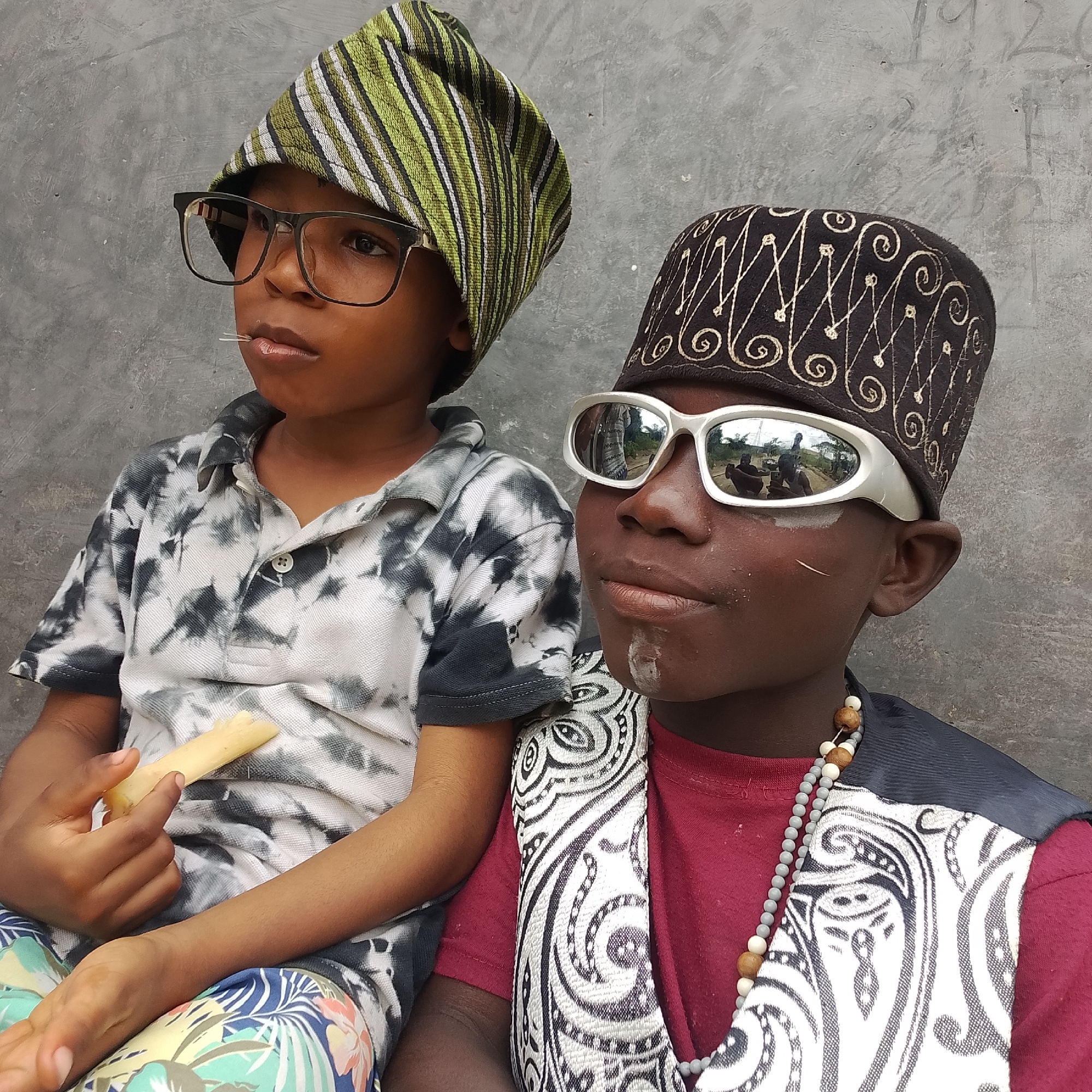 The image shows two young boys sitting in front of a gray concrete wall. The boy on the left is wearing a green and white striped hat, black-rimmed glasses, and a tie-dye t-shirt. He is holding a piece of food, possibly sugarcane, in his right hand, with a toothpick in his mouth. The boy on the right is wearing a black cap with intricate gold embroidery, white-framed sunglasses, a maroon t-shirt, and a vest with a black and white patterned design. He is also wearing a beaded necklace. White face paint is visible on his chin and cheek. The reflection in the sunglasses shows an outdoor scene with people and possibly some vegetation. The overall impression is a candid portrait of two children, likely in an urban environment. The location is stated to be Ado, Nigeria.