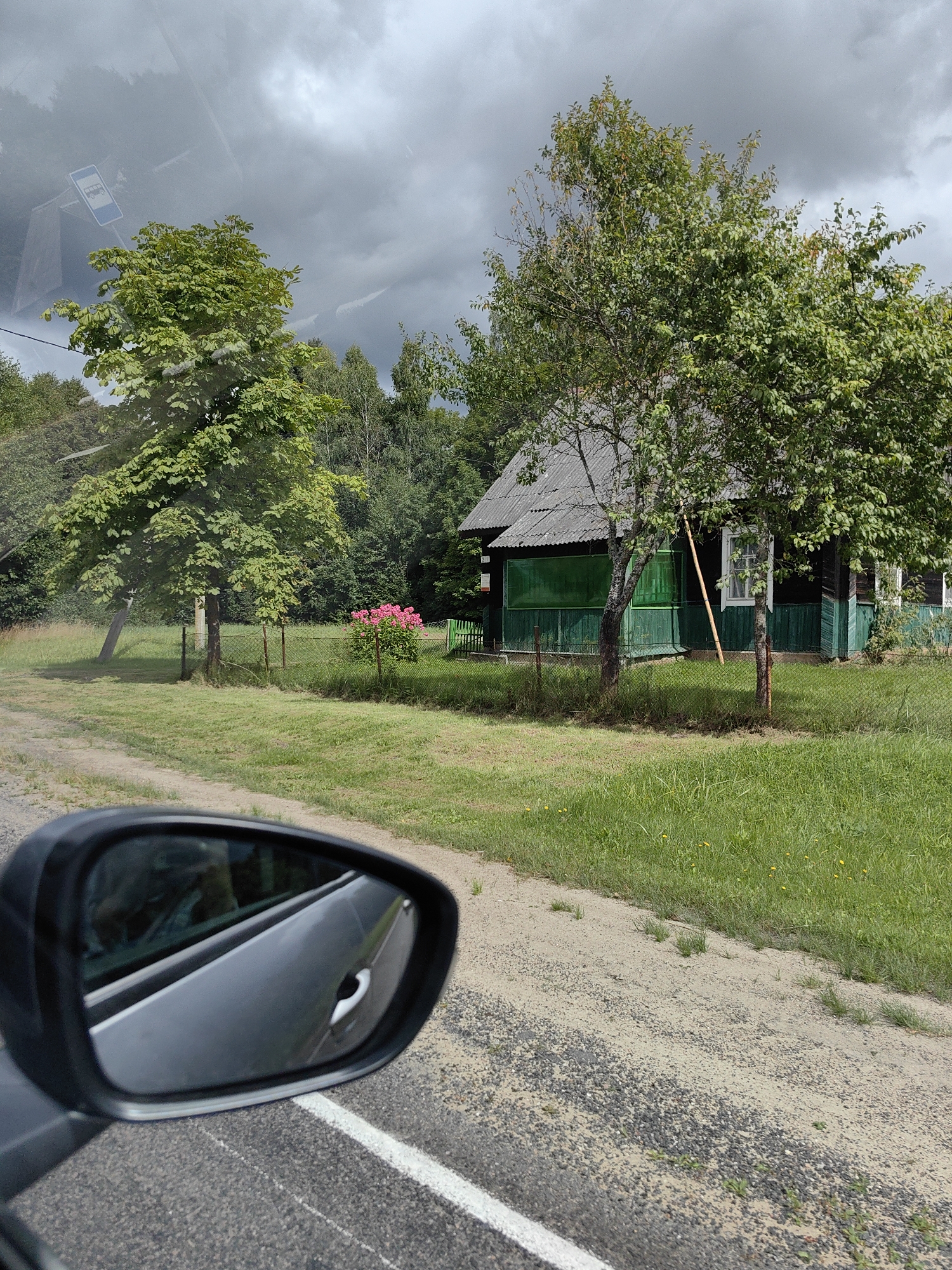 The image captures a rural roadside scene, likely in or near Мядзельскі сельскі Савет, Belarus, as suggested by the presence of a bus stop sign reflecting in the car's side mirror. In the foreground, the side mirror of a vehicle is visible, reflecting a blurred interior and the road ahead. The road itself is a gravel path with a white line marking its edge, bordering a grassy verge.
Dominating the midground is a dark wooden house with a gray, corrugated roof and green trim. A fence separates the property from the road. In front of the house, a vibrant cluster of pink flowers is in bloom. Several trees are scattered throughout the scene. A large deciduous tree with lush green leaves stands to the left of the house, while other trees, including one with a thinner trunk, are closer to the dwelling. The background is a dense forest under a cloudy, overcast sky. The overall impression is one of a quiet, rural settlement on a possibly inclement day.