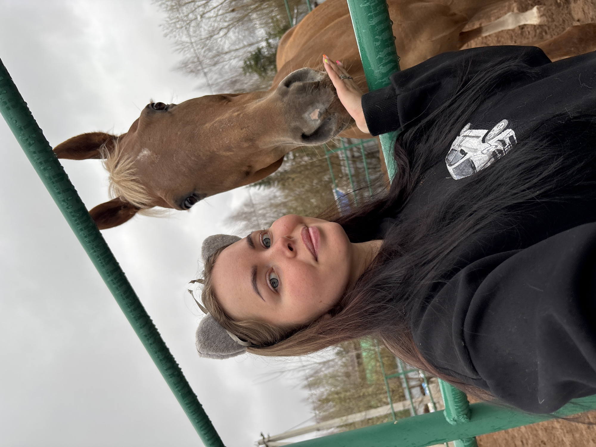 A young woman with light skin and blue eyes, wearing a black hooded sweatshirt and grey cat-ear headband, is interacting with a light brown horse with a blonde mane. The woman is holding the horse's muzzle with her right hand. They are standing near a green metal fence. The background includes trees and an overcast sky. The location is Русский Вожой, Russia.