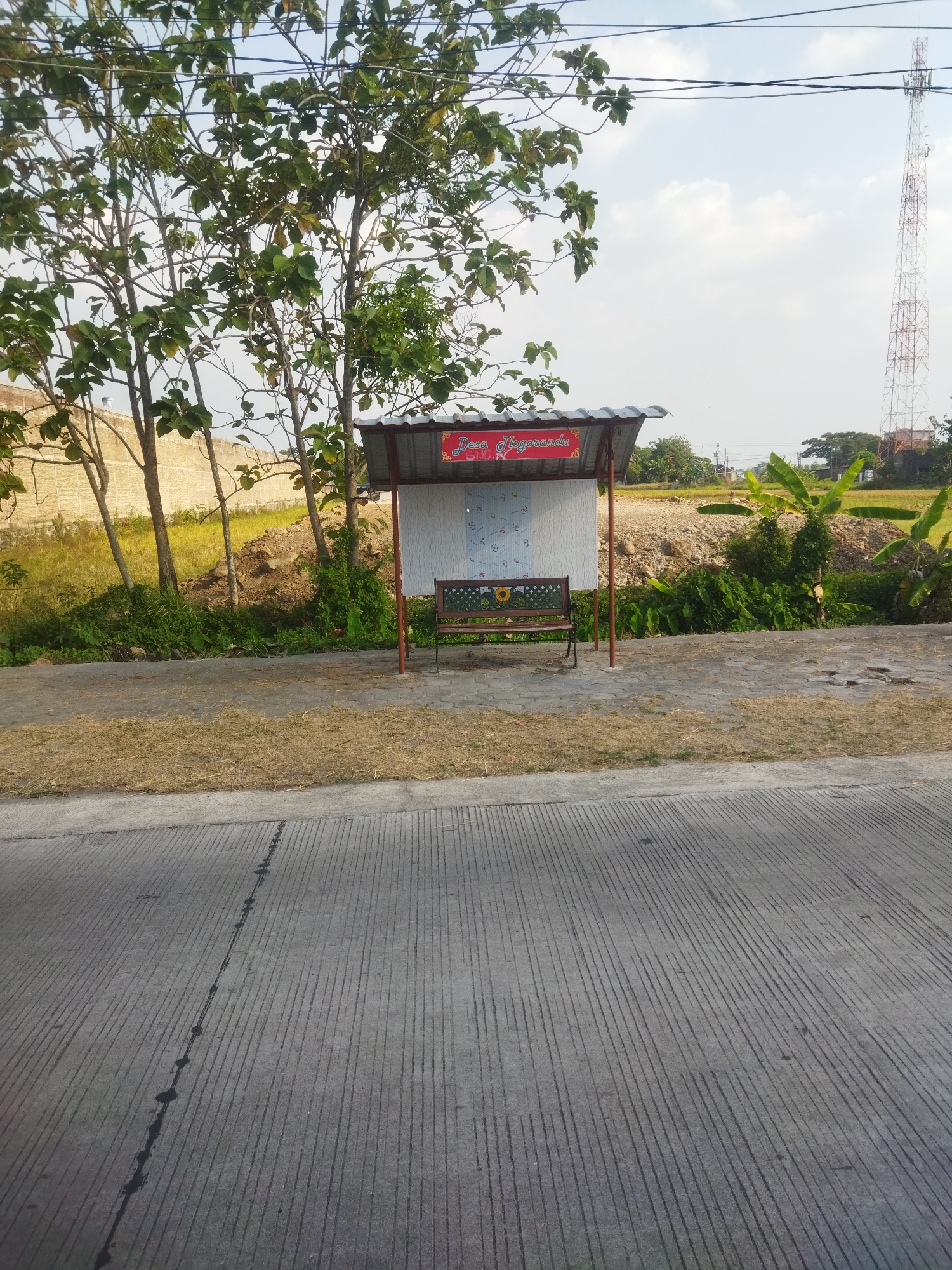 A shelter with a corrugated metal roof stands on a paved area, beside a road with a distinct striped pattern. A wooden bench with a metal frame and decorative sunflower motif is placed inside the shelter. Behind the shelter, a dry, grassy area transitions to a dirt embankment with some vegetation and trees. A tall, white and red telecommunications tower is visible in the distance, against a pale blue sky with scattered clouds. A sign above the bench reads "Desa Jlogorandu". The location is identified as Juwiring, Indonesia.