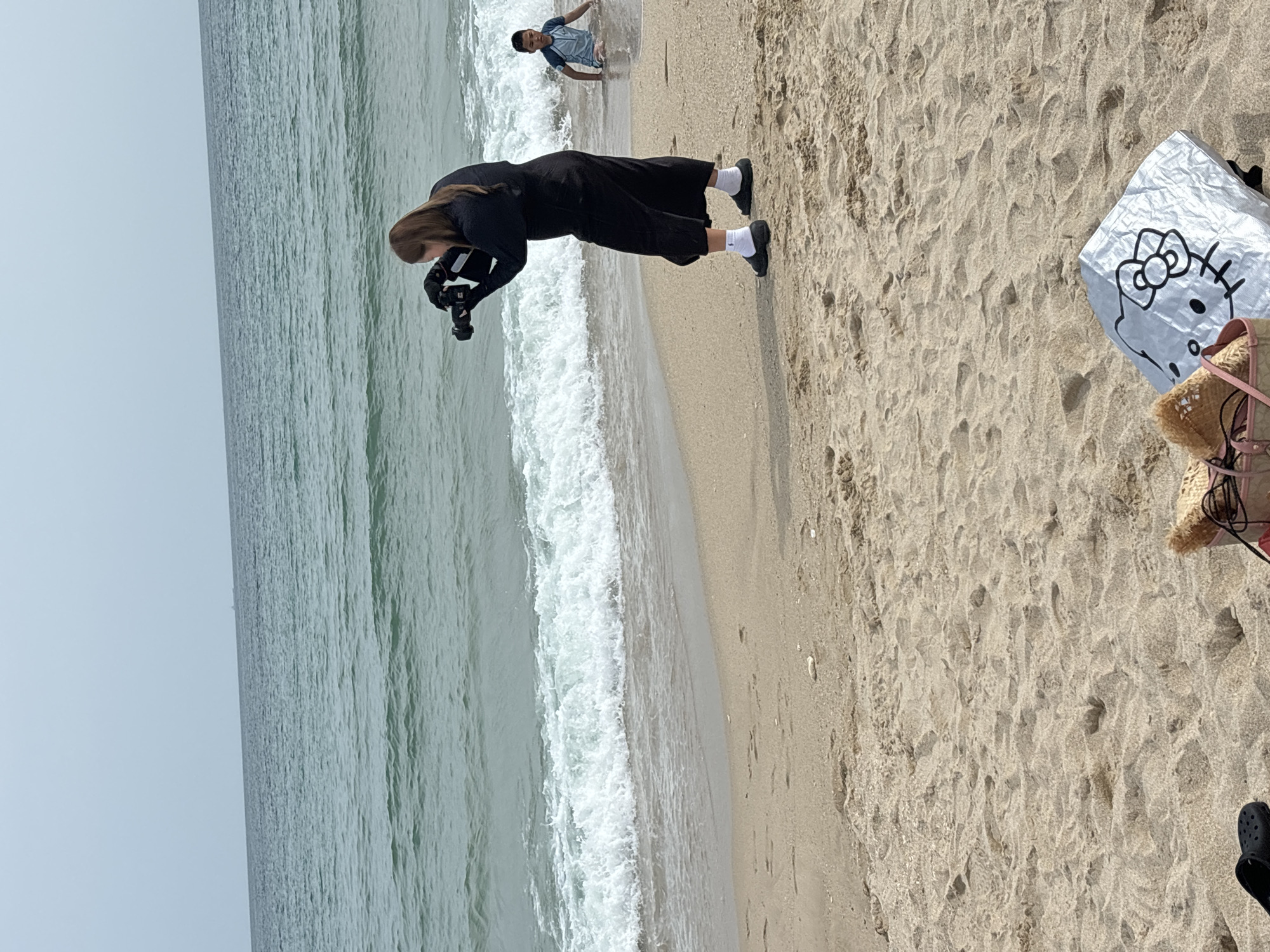 A woman in a black dress and white socks is taking a photo with a camera on a sandy beach. Next to her, a child in blue swim trunks is sitting in the shallow water. The ocean waves are breaking on the shore under a cloudy sky. In the foreground, a straw bag and a silver shopping bag with a Hello Kitty print are placed on the sand. The background shows a calm sea with a distant ship.