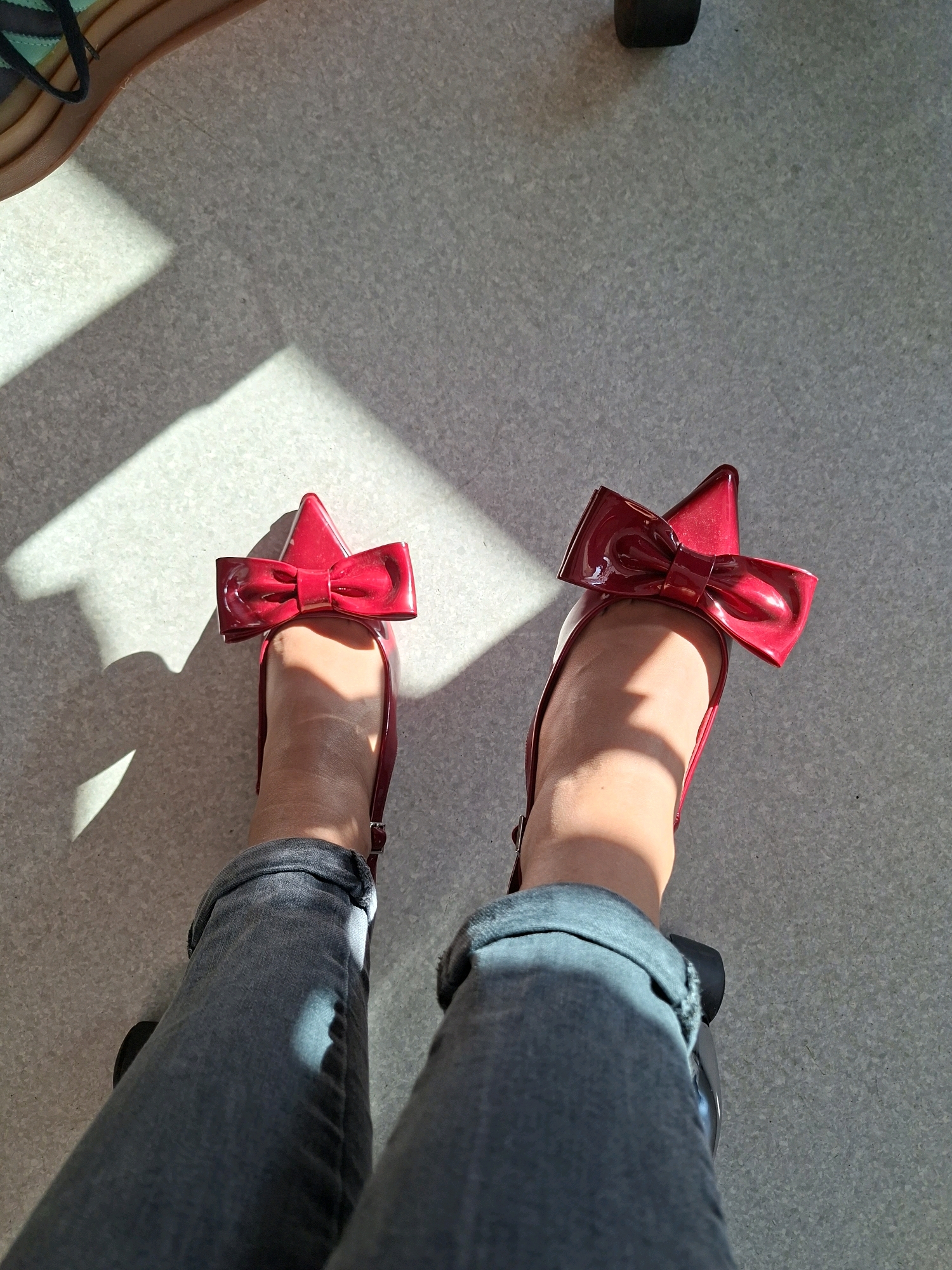 An overhead, eye-level medium shot shows a person's legs and feet, wearing red patent leather ballet flats with large red bows. The shoes are pointed with a thin strap across the heel. The person is wearing dark gray denim jeans that are cuffed at the ankles. The ground beneath is a light gray speckled surface, possibly concrete or linoleum. Sunlight streams in from the left, casting sharp shadows across the ground and the person's legs and feet. One shadow, likely from a window frame or railing, creates a strong rectangular pattern. Another shadow, softer and more diffuse, comes from the person's feet. The overall lighting suggests an indoor environment with natural light, possibly a sunroom, covered patio, or well-lit room. The focus is sharp on the shoes and the lower legs, with the background slightly softer.
