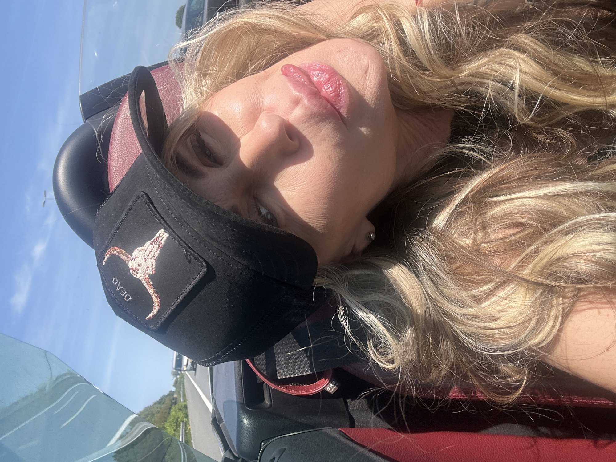 A woman with blonde, wavy hair is the focal point of the image, possibly in Leidschendam, Netherlands, though specific city landmarks are absent from the frame. She is wearing a black hat with a steer skull logo. The environment suggests she is in a vehicle with red leather seats and a windshield. The lighting is bright, indicating daytime.