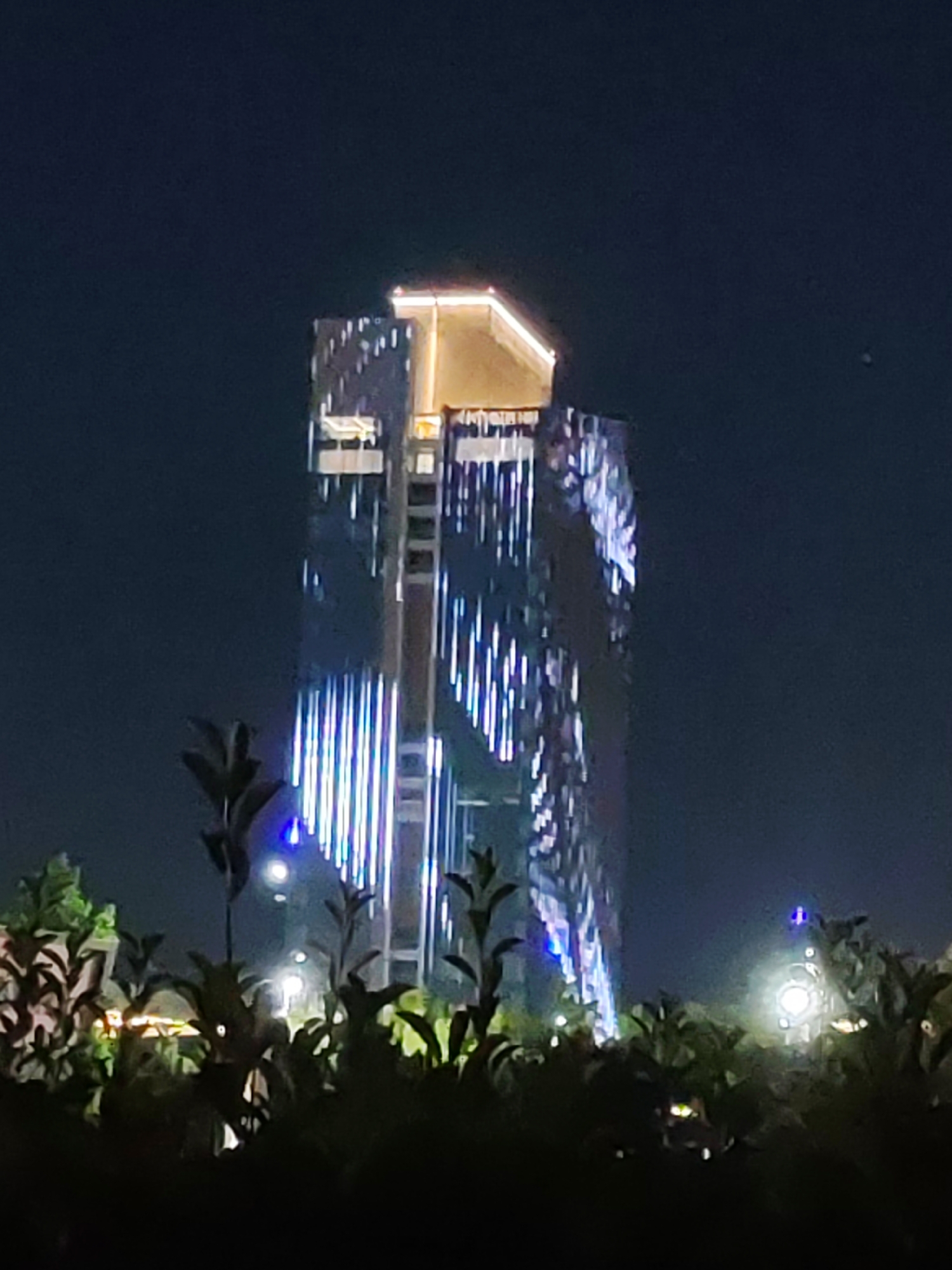 The image captures a nighttime view of a modern skyscraper in Tashkent, Uzbekistan. The building, characterized by its sleek, reflective facade, is illuminated with a dynamic light show. Vertical stripes of blue and white light cascade down its surfaces, creating a striking visual effect against the dark sky. The top of the skyscraper features a warm, golden light, highlighting its architectural design. In the foreground, silhouetted foliage and plants are visible, suggesting the photo was taken from a garden or park area. Several bright lights are scattered throughout the foreground, possibly streetlights or decorative elements, and a distant circular light source is also visible. The overall scene conveys a sense of urban sophistication and vibrant nightlife.