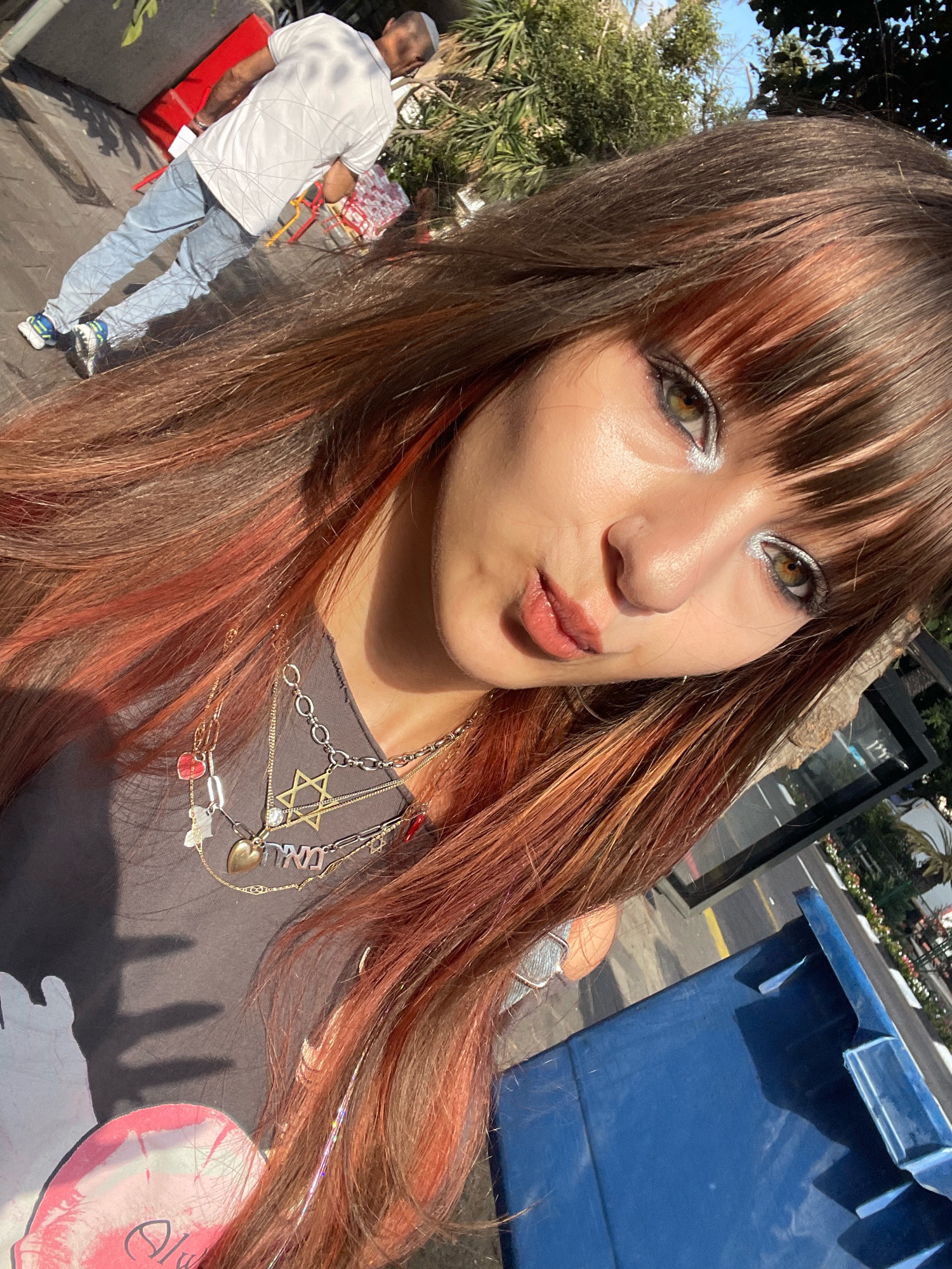 In Raanana, Israel, a young woman with reddish-brown hair and bangs takes a selfie outdoors. She is wearing a grey t-shirt layered with multiple necklaces, including a Star of David pendant, a heart pendant, and several charms. Her makeup includes silver eyeshadow. In the background, a man in a white shirt and blue jeans stands, looking down. There are red and white shopping bags near him, and a blue dumpster is visible to the right. The scene appears to be on a street or sidewalk with vegetation and shadows.