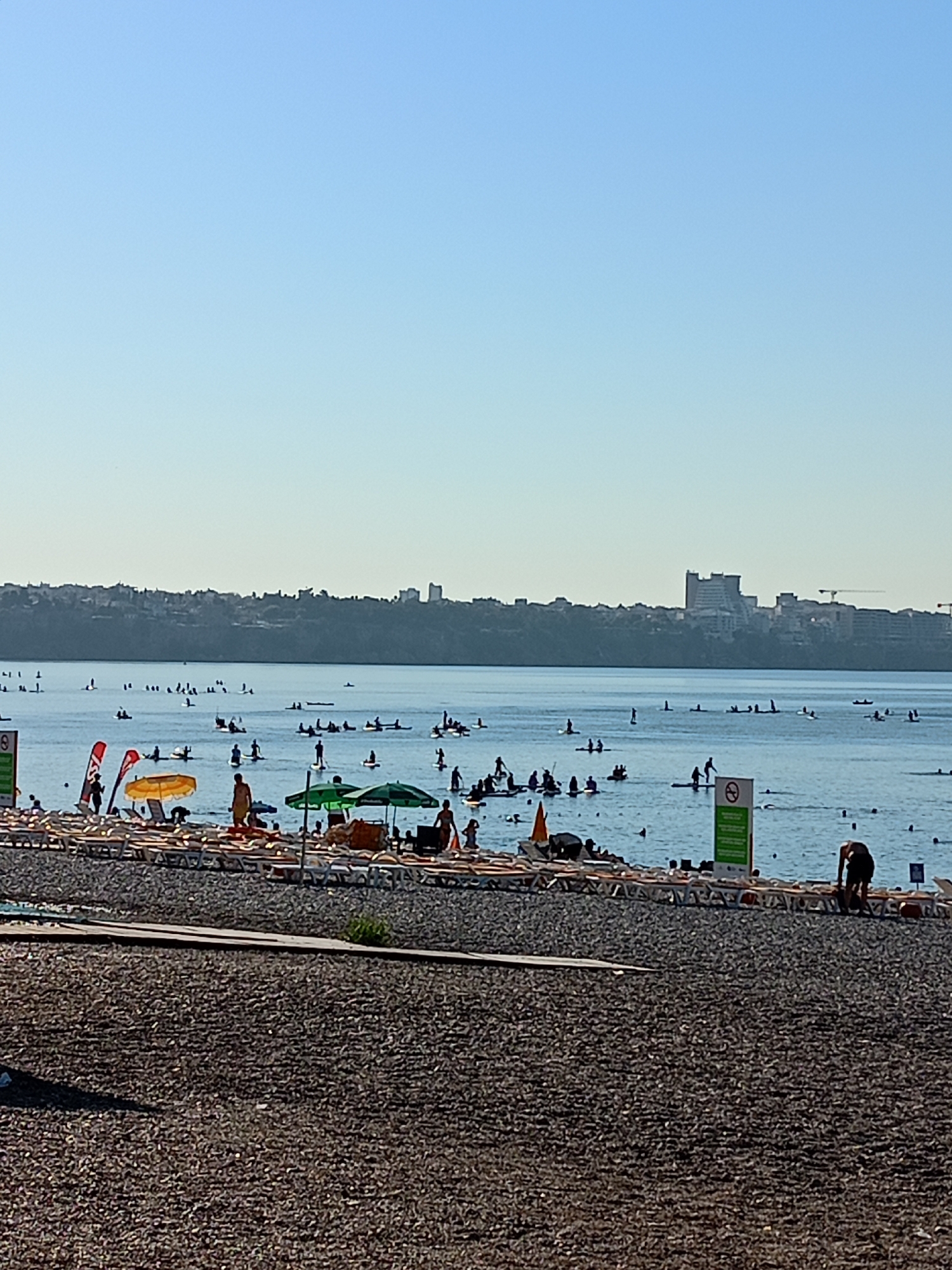 This image depicts a sunny day at a beach in Antalya, Turkey. The foreground is a pebble beach with rows of empty lounge chairs. In the middle ground, the calm blue sea is dotted with numerous people engaged in various water activities, primarily stand-up paddleboarding. Several individuals are on paddleboards, while others are swimming. Two green beach umbrellas and one yellow umbrella are visible on the beach, providing shade. In the background, a city skyline with buildings and cranes lines the horizon, characteristic of Antalya's urban coastal landscape. The sky is a clear, bright blue, indicating fair weather.