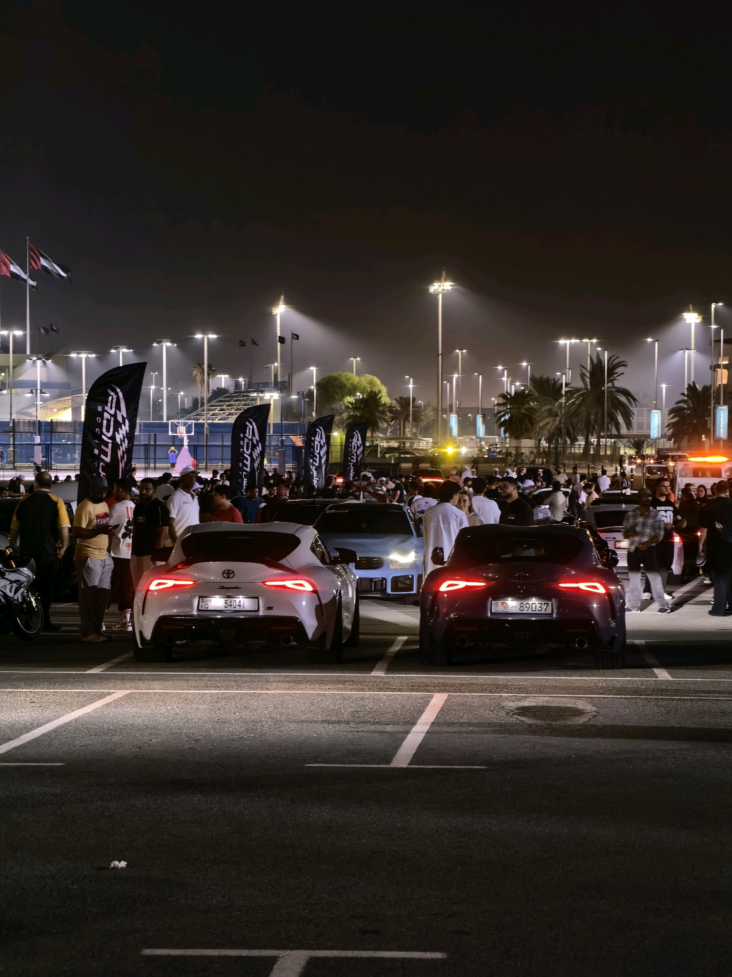 The image captures a nighttime scene in Abu Dhabi, United Arab Emirates, likely at an outdoor event or gathering. The foreground is dominated by two sports cars, a white Toyota Supra on the left and a dark red Toyota Supra on the right, parked on an asphalt surface marked with white parking lines. Both cars have their brake lights illuminated, suggesting they are stationary. The white Supra displays a license plate reading "54041". The red Supra has a license plate reading "89037".
A crowd of people is gathered around the cars and in the background, illuminated by numerous bright streetlights and the headlights of vehicles. Many individuals appear to be conversing and observing the cars. Several large black flags with white lettering, possibly representing a brand or organization, are prominently displayed among the crowd. In the far background, palm trees are visible, and the silhouette of a stadium or sports facility with lighting can be discerned. The overall atmosphere appears to be that of a car show or a social event.