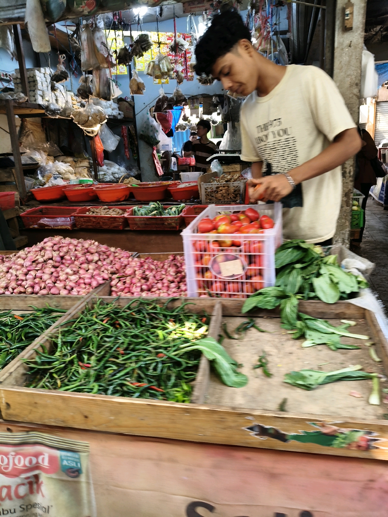 A young man wearing a light-colored t-shirt with black text is standing at a market stall in Setiamekar, Indonesia. He is holding a red tomato in his right hand and appears to be sorting or preparing it. In front of him, to his left, is a large white crate filled with ripe red tomatoes. To the right of the tomatoes is a pile of leafy green vegetables. Further to the left and in the foreground, there is a wooden crate overflowing with long green chilies. Behind the chilies, to the left, is a large mound of red shallots. The market stall is cluttered with various items hanging from the ceiling, including bags of goods and what appear to be dried or preserved foods. In the background, other people can be seen at different stalls. The overall environment suggests a bustling open-air market.