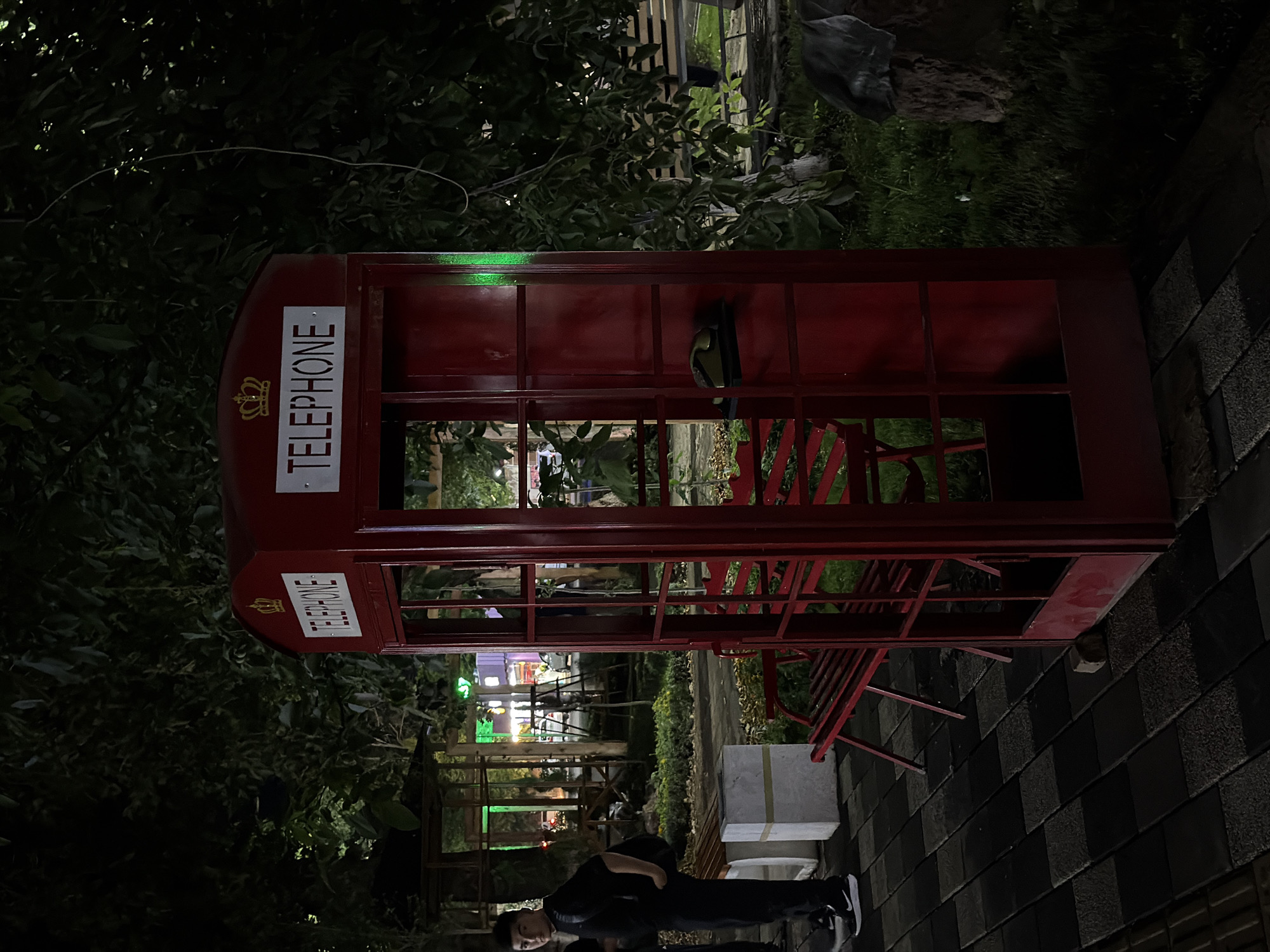 The image depicts a red British-style telephone booth situated in an outdoor urban setting at night. The booth has the word "TELEPHONE" written in white lettering above a crown emblem. Inside the booth, a black telephone receiver rests on a shelf. The surrounding environment features trees with dark foliage, benches, and a paved walkway. In the background, illuminated buildings and structures suggest a city environment. A young person in dark clothing stands to the left of the telephone booth, facing away from the camera. The scene is lit by artificial lights, casting shadows and creating a nighttime ambiance. This image was taken in the city of Chirchik, Uzbekistan.