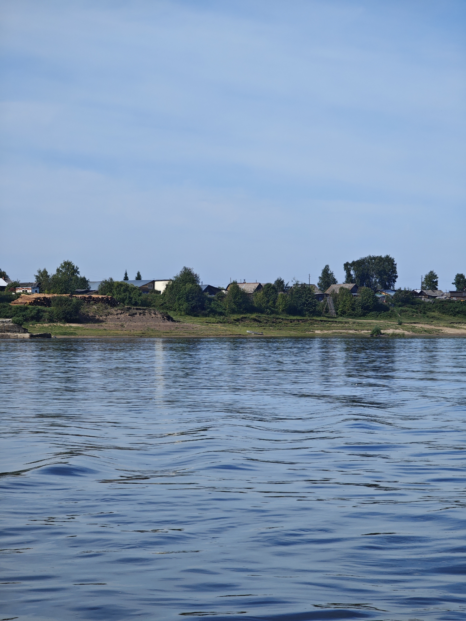The image depicts a wide, calm body of water in the foreground, with gentle ripples reflecting the blue sky. In the background, a village is situated on a gentle slope leading down to the water's edge. The village consists of several small houses with pitched roofs, nestled amongst lush green trees. A pile of logs is visible on the bank, suggesting some form of industry or construction. The sky above is a clear, pale blue with faint wisps of clouds. The scene conveys a sense of rural tranquility. The location is stated to be the city of Yartsevo, Russia.