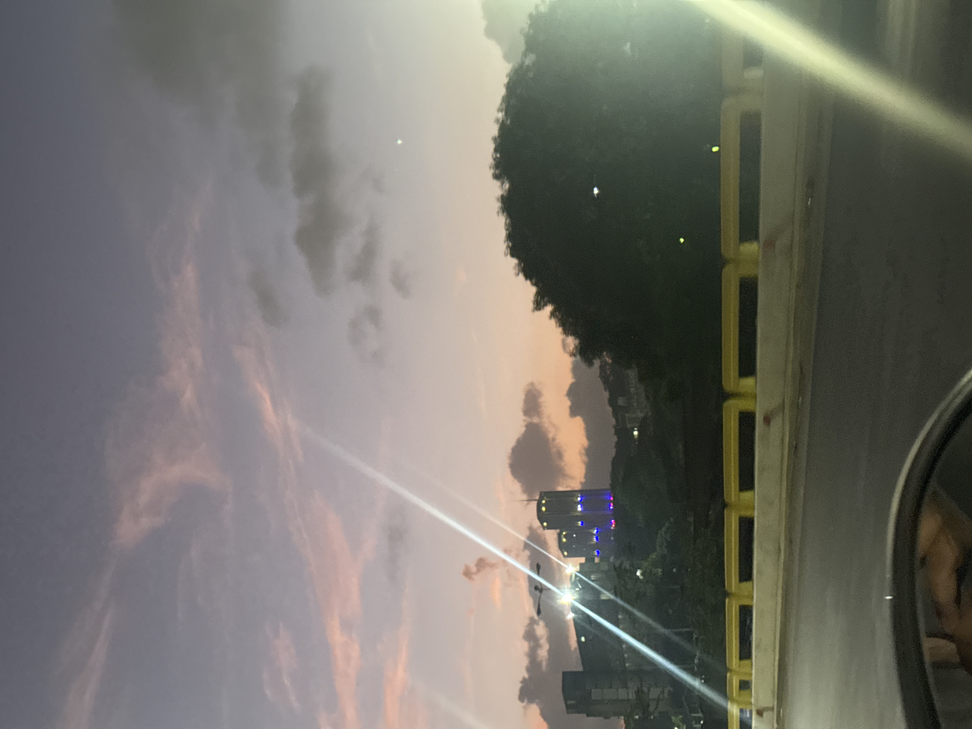 The image captures an urban twilight scene, likely viewed from an elevated position such as a bridge or overpass. The sky, dominating the upper half of the frame, displays the soft hues of sunset or dusk, transitioning from pale orange and pink near the horizon to shades of purple and grey in the upper reaches, dotted with scattered clouds.
On the right side, a dense line of dark green foliage or trees suggests a wooded area or park within the city. Below the horizon, numerous urban buildings are visible in the distance, some with illuminated windows, indicating the onset of night. One prominent building in the midground features distinct blue and purple accent lighting on its facade.
In the immediate foreground on the right, a distinctive yellow and grey concrete railing or barrier runs horizontally, separating the viewpoint from the landscape below. To the far right, a portion of a vehicle's side mirror is visible, reflecting a distorted image of what appears to be part of the car's interior or possibly a person inside. Bright, elongated streaks of light, likely lens flares from strong artificial light sources such as streetlights or car headlights, cut diagonally across the frame, adding a dynamic element to the image. The overall impression is that of a quiet moment capturing the transition from day to night over a city like Caracas, with no discernible human activity or visible text.
