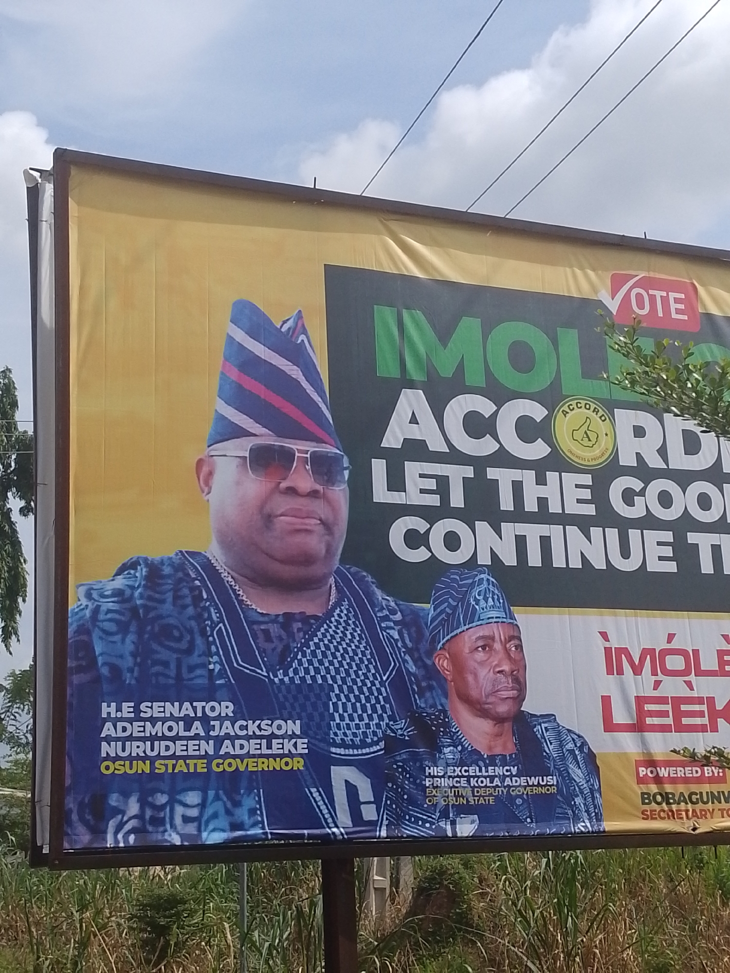 A billboard on a pole displays two men in traditional Nigerian attire. The man on the left is wearing a blue and white patterned outfit and a blue, red, and white striped cap. He has sunglasses and a silver chain. The man on the right is wearing a dark blue patterned outfit and a blue patterned cap. The billboard has text that reads "VOTE", "IMOLE", "ACCORD", "LET THE GOOD CONTINUE TH", "H.E SENATOR ADEMOLA JACKSON NURUDEEN ADELEKE OSUN STATE GOVERNOR", "HIS EXCELLENCY PRINCE KOLA ADEWUSI EXECUTIVE DEPUTY GOVERNOR OF OSUN STATE", and "POWERED BY: BOBAGUNWA SECRETARY TO". The background is yellow and green. The billboard is outdoors, with green foliage and tall grass visible at the bottom, and wires and a cloudy sky above. The image was taken in Abeere, Nigeria.