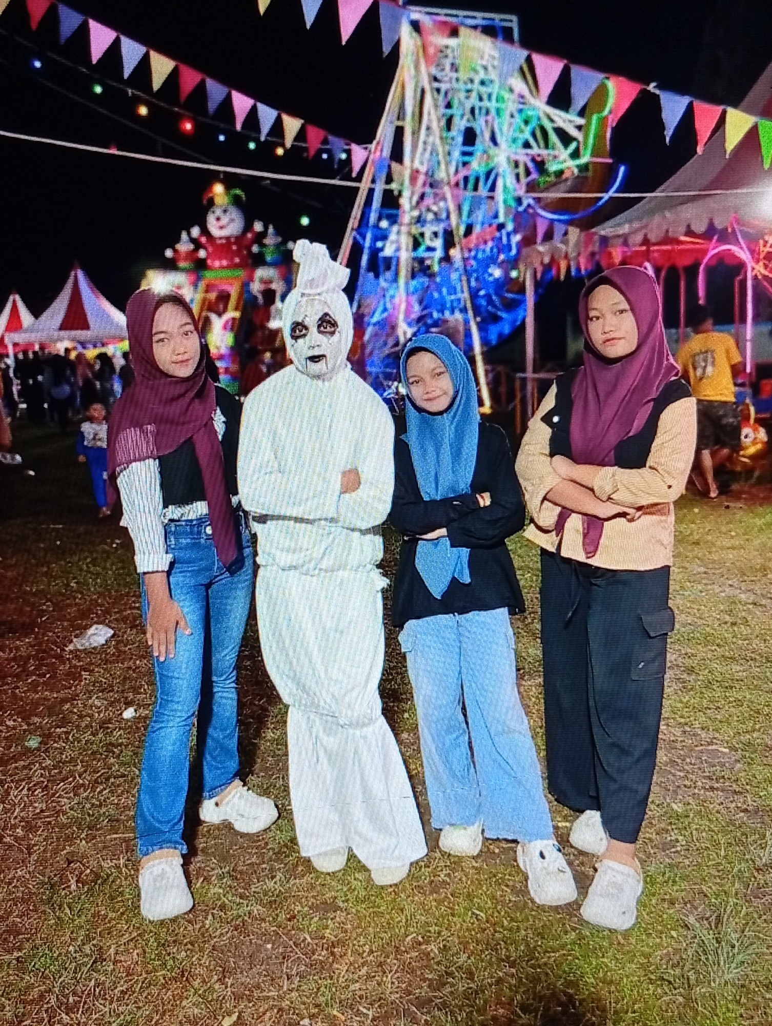 This image, taken at a night fair or carnival in Lhokseumawe, Indonesia, features four individuals posing in the foreground against a brightly lit background. The ground is a dark, patchy mix of dirt and sparse grass, with some discarded white debris visible in the immediate foreground.
From left to right, the subjects are:
1. A young female wearing a dark maroon hijab, a long-sleeved striped shirt layered under a black top, and blue denim jeans. She wears white platform clogs and has a slight smile, with her left hand resting at her side.
2. A person in a full-body white costume, featuring a mask-like face with prominent dark eye markings and a painted mouth, along with a white head covering. This individual stands with arms crossed over the chest, wearing white footwear.
3. A young female wearing a light blue hijab, a black long-sleeved top, and light blue wide-leg trousers. Her arms are crossed over her chest, and she wears white platform clogs, displaying a faint smile.
4. A young female wearing a dark maroon hijab, a two-tone top (light beige or yellow long-sleeved shirt with a black overlay), and dark-colored cargo pants. Her arms are crossed over her chest, and she wears white platform clogs, maintaining a neutral expression.
In the mid-ground and background, a large, multi-colored illuminated Ferris wheel is prominent on the right. To its left, another illuminated structure resembling a clown or character, primarily red and green, is visible. Multiple red and white striped tents are dispersed throughout the background, indicating various stalls or attractions. Colorful triangular pennants are strung horizontally across the upper portion of the frame, further enhancing the festive atmosphere. Other indistinct figures are present in the background, including a child in blue behind the leftmost subject and an adult male in a yellow t-shirt further back on the right.