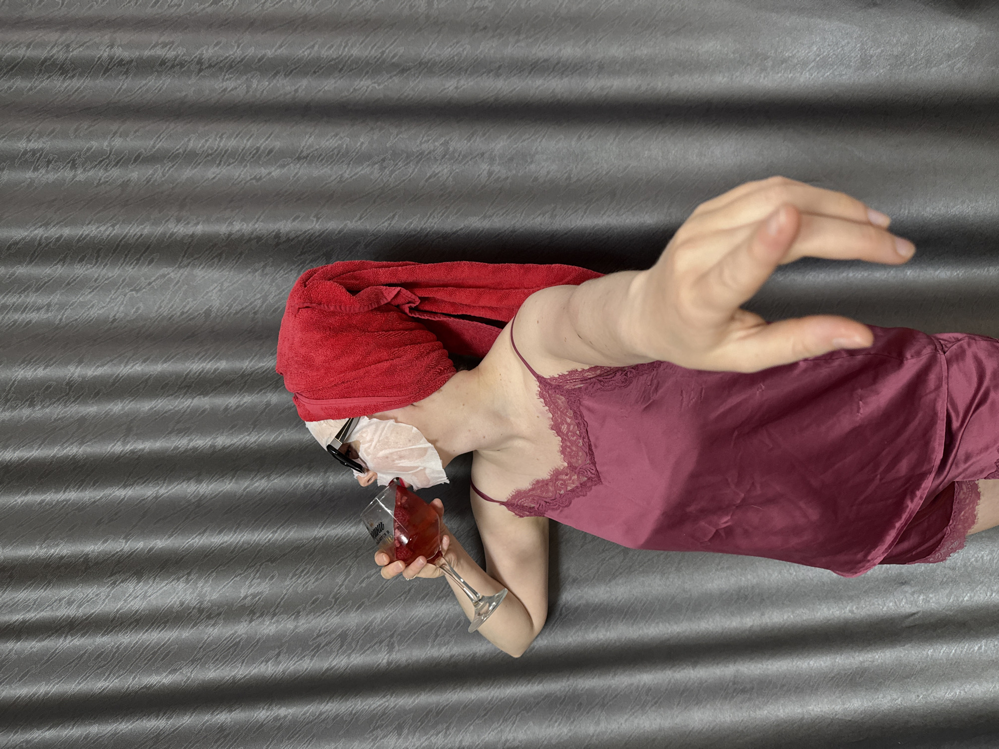 A woman is lying on a gray, textured surface. She has a red towel wrapped around her head, a white facial mask on, and glasses perched on the mask. She is wearing a maroon nightgown with lace trim and has a glass of red liquid in her left hand. Her right arm is extended away from her body. The location and city are unknown.