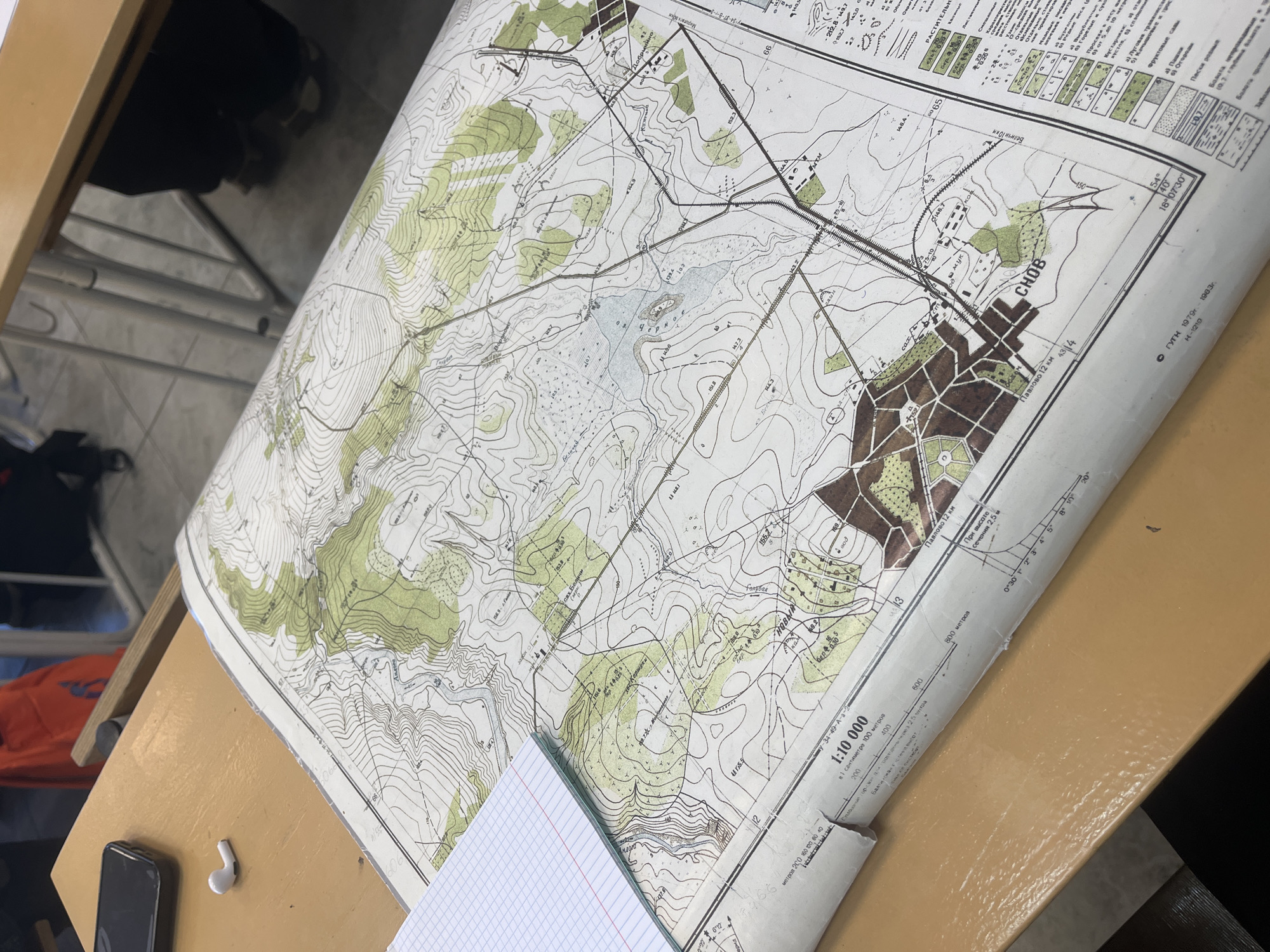 The image shows a topographical map laid out on a table. The map is printed in several colors and shows contour lines, vegetation, roads, and other features. The bottom of the map indicates a scale of 1:10,000 and the name "CHOB" and "Павлово 12 км". The map also says "54°40 / 18°07'30"." The map legend shows various symbols, including types of vegetation, terrain, and structures. A cell phone, an earbud, and a notepad are placed on the table near the map. The scene is indoors, likely in a classroom or office setting, as indicated by the table and nearby objects. Based on the text "Павлово 12 км" the location of this topographical map is Павлово, Saint-Petersburg, Russia.