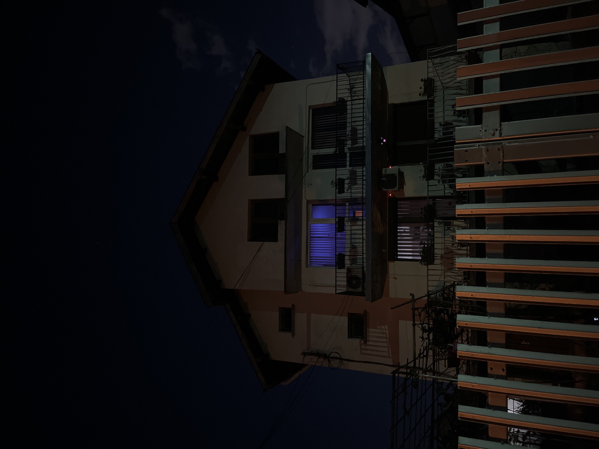 The image captures a nighttime scene of a multi-story building in an urban environment. The photograph appears to be rotated 90 degrees clockwise; when viewed upright, the dark sky with subtle cloud formations occupies the upper portion, while the building fills the central and left-leaning areas.
The main subject is a building with light-colored walls, possibly stucco or concrete, and a prominent, steeply pitched roofline on its left side. Numerous windows are visible across its facade; one on a middle floor stands out, emitting a distinct bright blue light. Other windows appear dark or have closed blinds/shutters. Several air conditioning units are mounted on the exterior walls, with small red indicator lights visible on some. A black metal structure, likely a fire escape or an external staircase, runs vertically down the right side of the building's main section.
On the far right, a large, dark, slatted structure, possibly a fence, balcony screen, or part of an adjacent building, partially obscures the view. Its horizontal slats are dark and metallic or wooden. The overall lighting is dim, characteristic of night, with the blue window light providing a significant contrast.
There are no people or discernible activities taking place. No visible text is present in the image. The scene conveys a quiet, static urban nightscape, located in Sofia, Bulgaria.