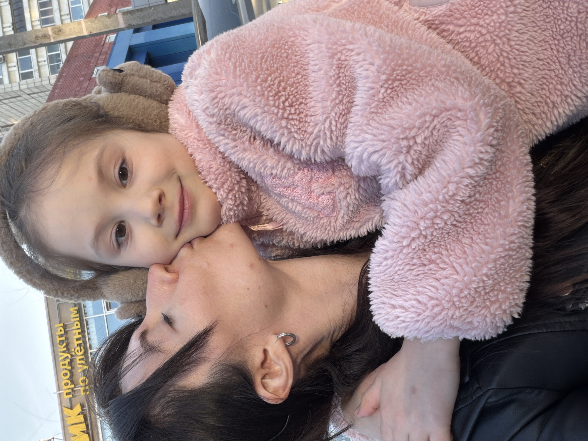 A close-up, somewhat tilted shot features a young girl wearing a pink fleece jacket and furry earmuffs leaning against an adult woman. The girl smiles and looks directly into the camera. The woman has dark hair, fair skin, and an earring in her left ear. She has her eyes closed and appears to be embracing the girl.
In the background, a building with windows is partially visible. A sign with yellow and black lettering that reads "Продукты по улетным" can also be seen. Based on the presence of the Russian text, this photo was taken in a Russian-speaking location. Specifically, the sign "Продукты по улетным ценам" is from a branch of the supermarket "Semya" in the city of Perm, Russia.