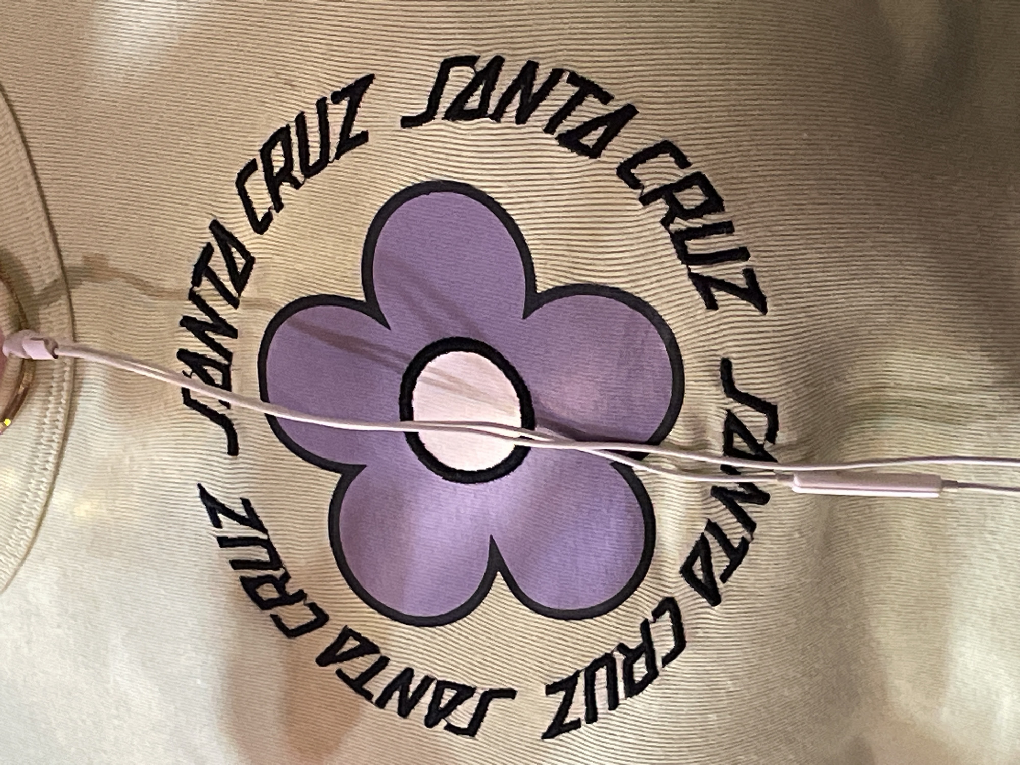The image shows a close-up of a light green or beige fabric, likely a t-shirt, with a circular design printed on it. The design features a purple flower with a black outline and a white center. Encircling the flower in a ring is the phrase "SANTA CRUZ" in black, blocky lettering. A pair of light pink headphones or earbuds are draped across the image, obscuring parts of the design.
The scene does not provide enough information to determine a location in L'Hospitalet De Llobregat, Spain. There is no information about people, subjects, objects, actions, interactions, or the environment.