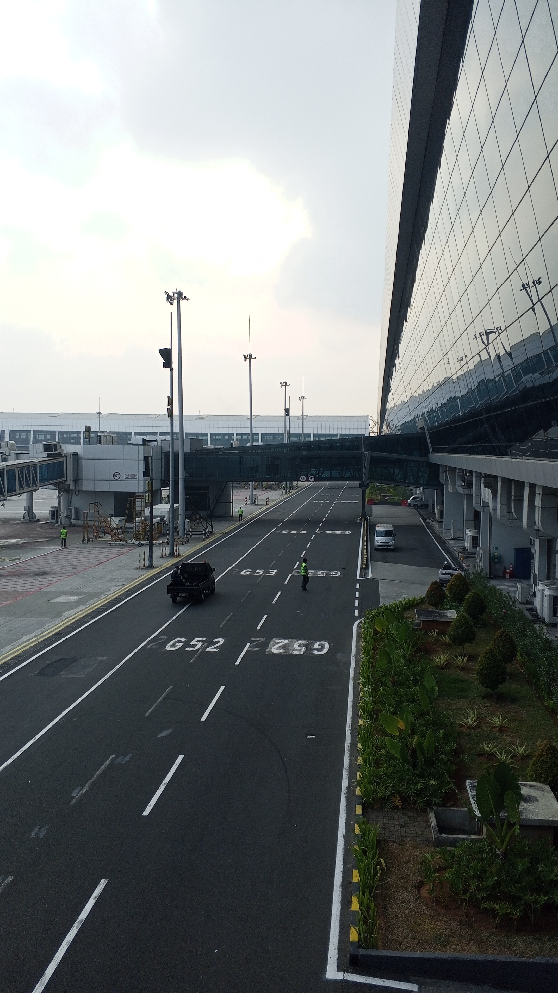 The image captures a scene at an airport in Rawa Burung, Indonesia. A modern airport terminal building with a glass facade dominates the right side of the frame. To the left, a jet bridge extends from the terminal to an aircraft, and beyond that, another section of the airport infrastructure is visible, including a long, enclosed walkway connecting different parts of the terminal.
The foreground features a paved road with markings indicating aircraft taxiing areas, labeled "G52" and "G59". A black pickup truck is driving away from the viewer on this road. Several individuals wearing high-visibility yellow vests are present on the tarmac, seemingly engaged in airport operations. One individual is standing near the pickup truck, while another is further down the road.
The environment appears to be an outdoor, functional area of an airport. The sky is overcast with bright clouds, suggesting daylight hours. A strip of greenery with bushes and trees lines the edge of the paved area on the right side of the road. The overall impression is one of a busy, operational airport environment.