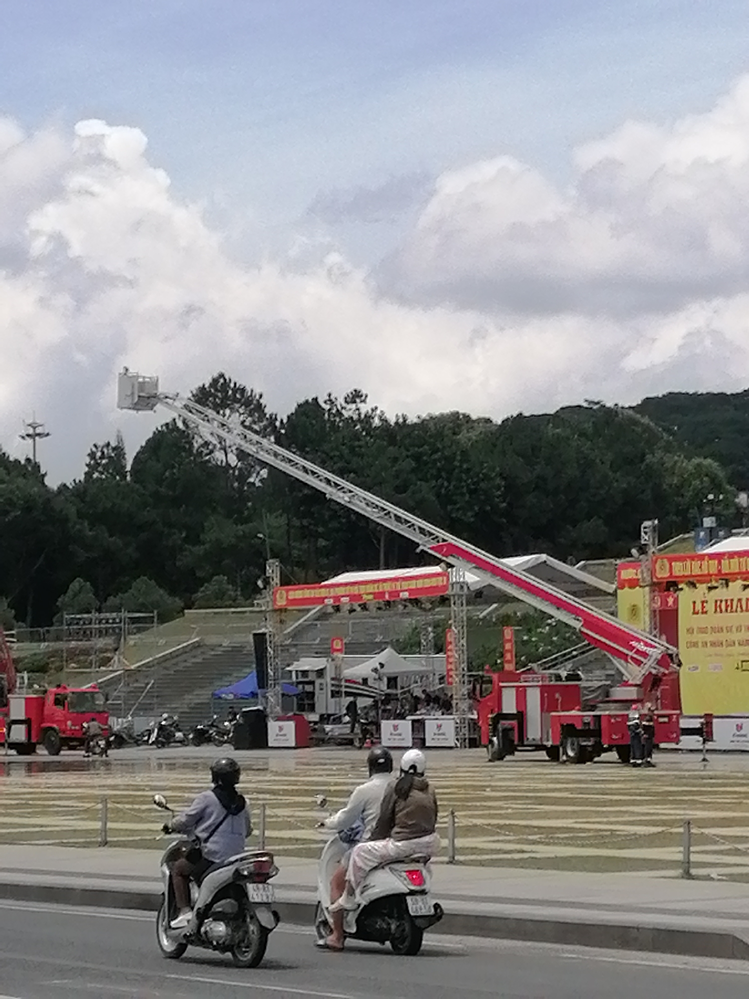 The image shows a wide outdoor space in Da Lat, Vietnam, likely a plaza or park, with a large crowd gathered in the background. A prominent fire truck with a long, extendable ladder is positioned in the middle ground. The ladder is angled upwards, with a basket at the end, suggesting it is part of a demonstration or event. Several smaller vehicles, possibly scooters, are visible in the foreground, with two people riding on one. The background features trees, hills, and a cloudy sky. A large banner with Vietnamese text is displayed on a yellow structure, hinting at a local event or festival. The scene suggests a public gathering with a focus on emergency services or a similar demonstration.